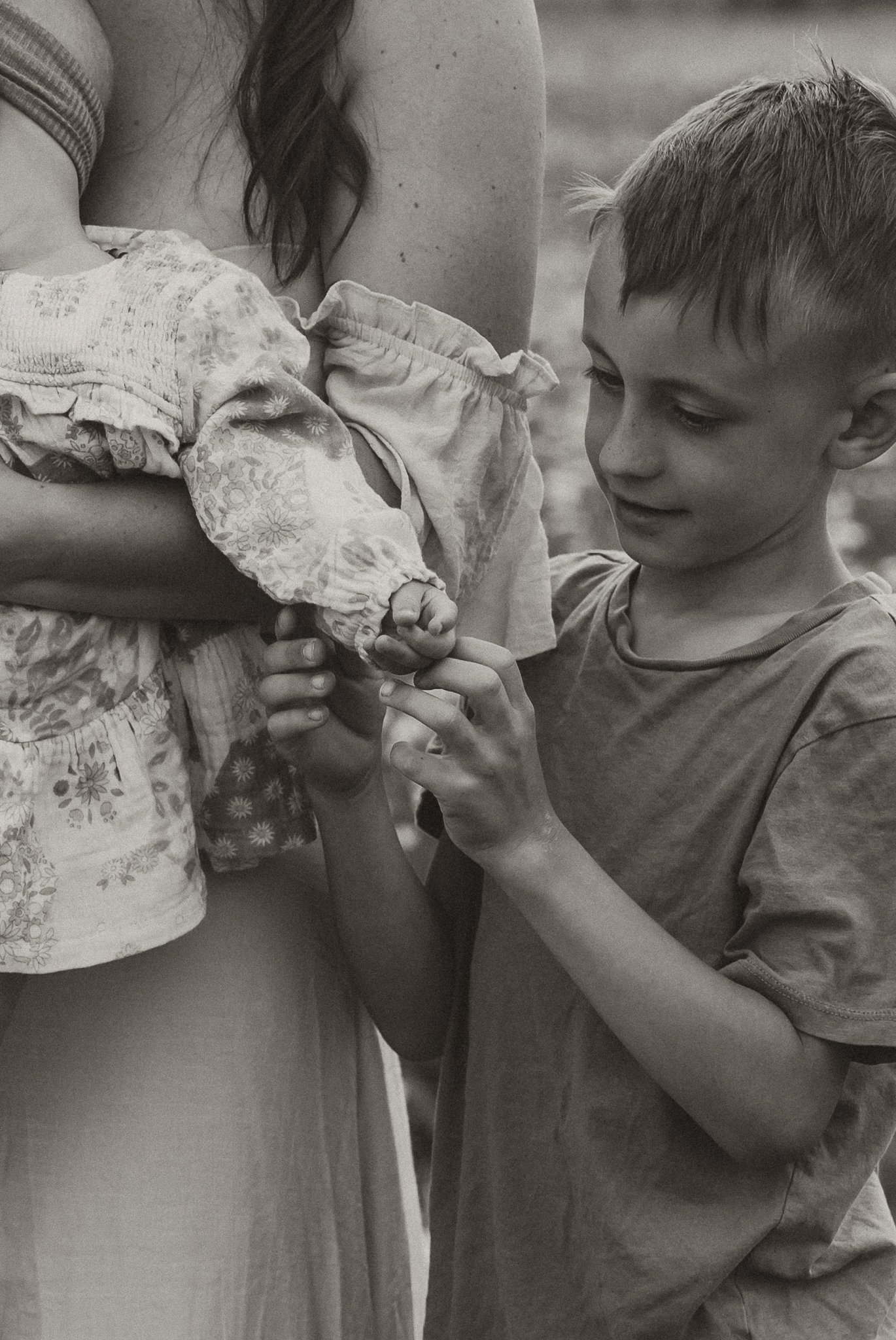 A young boy with short hair holding and looking at a small baby girl, who is being held by an adult woman. The image is in black and white.