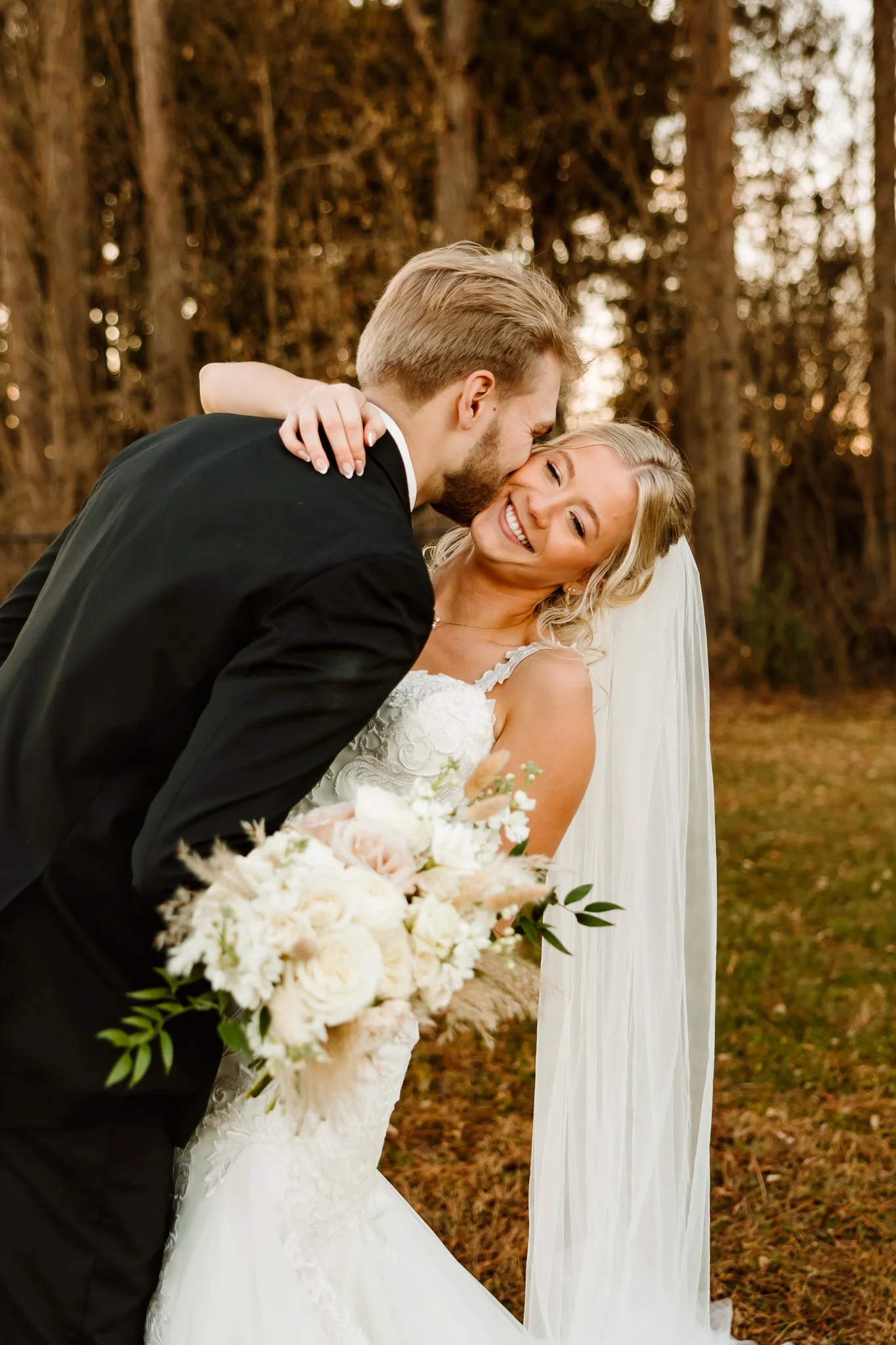 A newlywed couple sharing a joyful moment outside, with the groom leaning in to kiss the bride's forehead, she smiling joyfully, holding a bouquet of white and blush flowers, with trees in the background.  Cambridge Minnesota 
