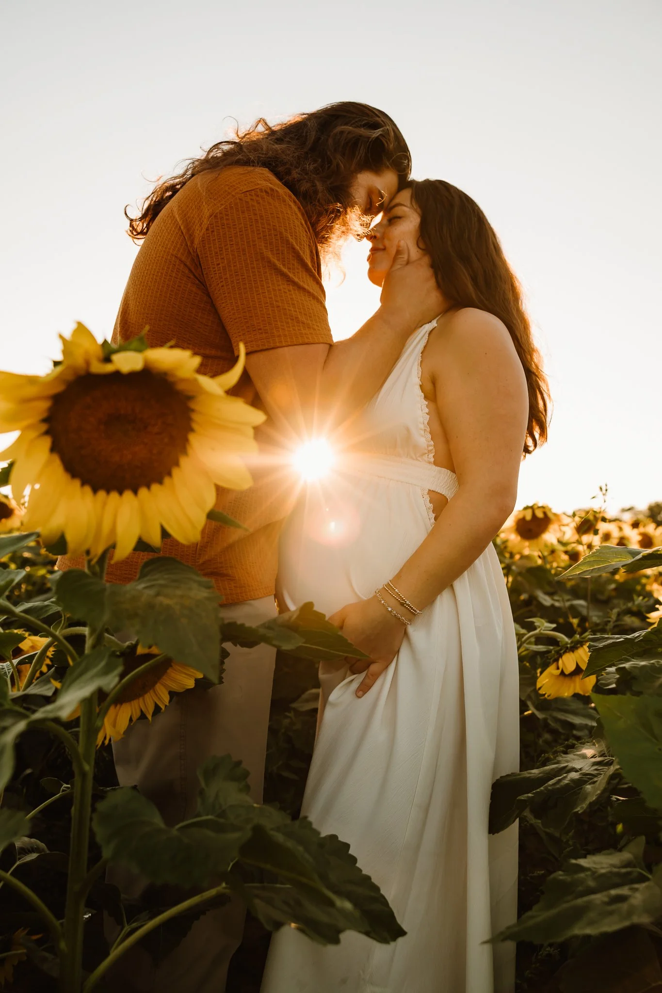 A couple standing close together in a sunflower field during sunset, touching foreheads, with the sun behind them creating a starburst effect.