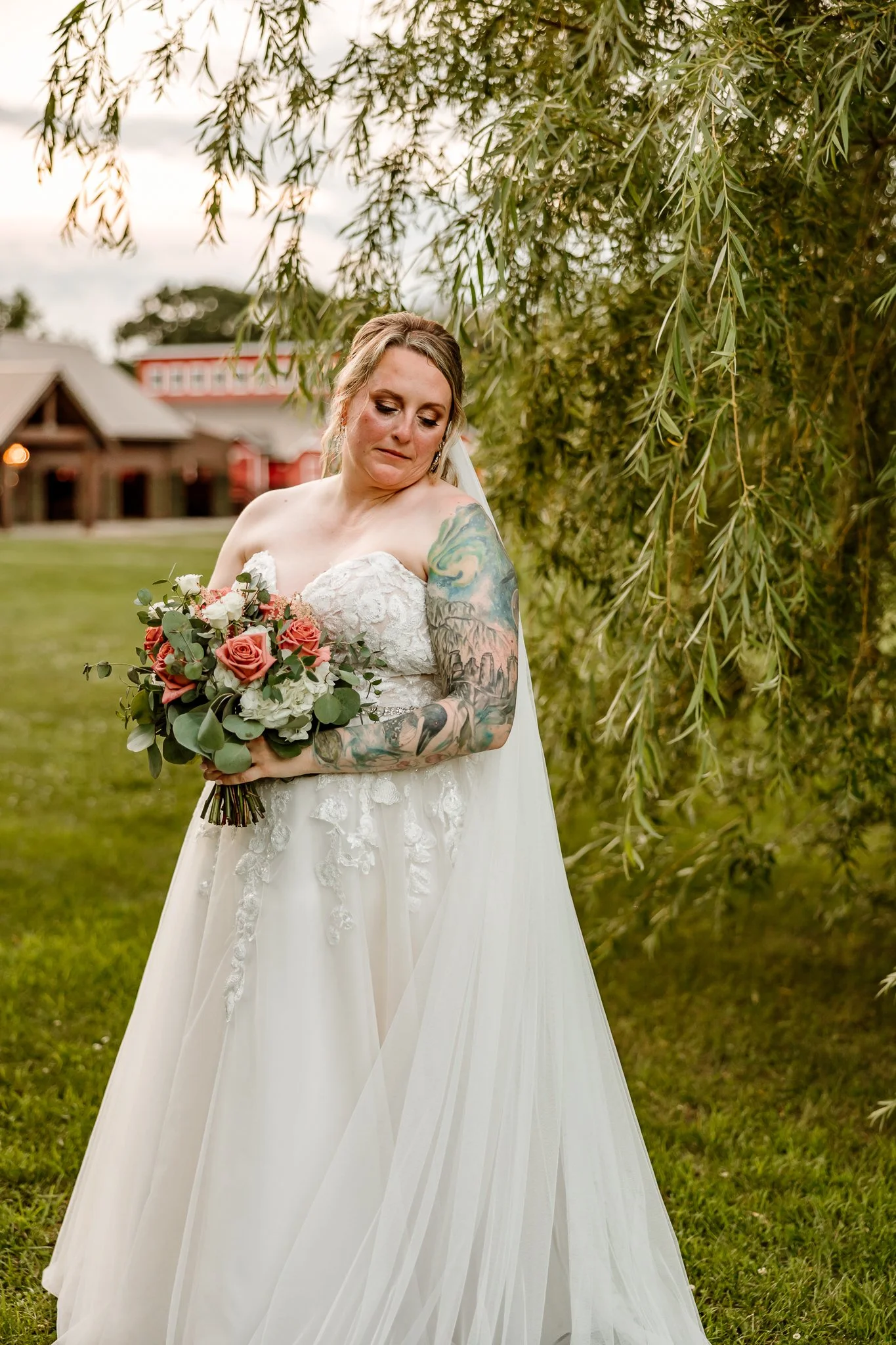 Bride in a strapless white wedding dress holding a bouquet of pink and white roses, standing outdoors near a leafy tree with a rustic building in the background.