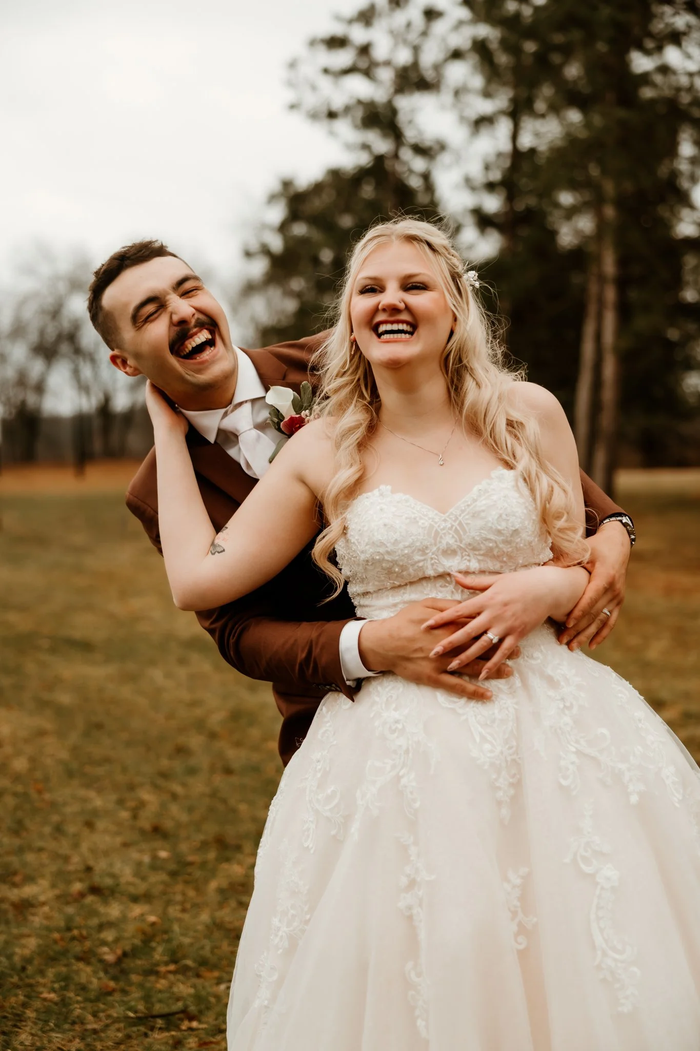 A bride and groom laughing and smiling outdoors during their wedding, with the groom holding the bride around her waist.