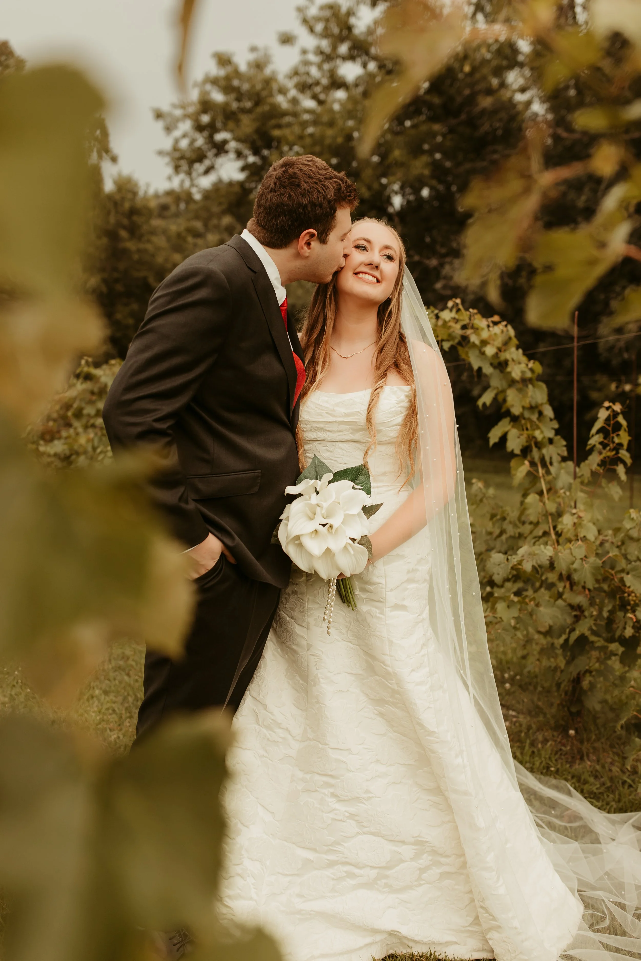A newlywed couple outdoors, with the groom kissing the bride on the cheek. The bride is smiling, holding a bouquet of white calla lilies, wearing a white wedding gown and veil. The groom is dressed in a black suit with a red tie. The background featu