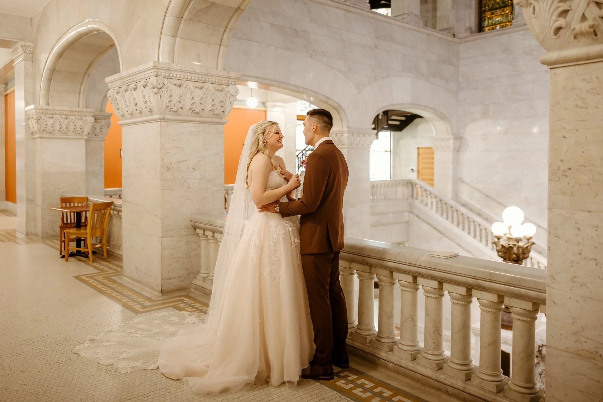 A bride and groom standing close together on a marble balcony inside a grand, historic building. The bride is in a white wedding gown with a long veil, and the groom is in a brown suit. They are smiling at each other in a romantic moment.