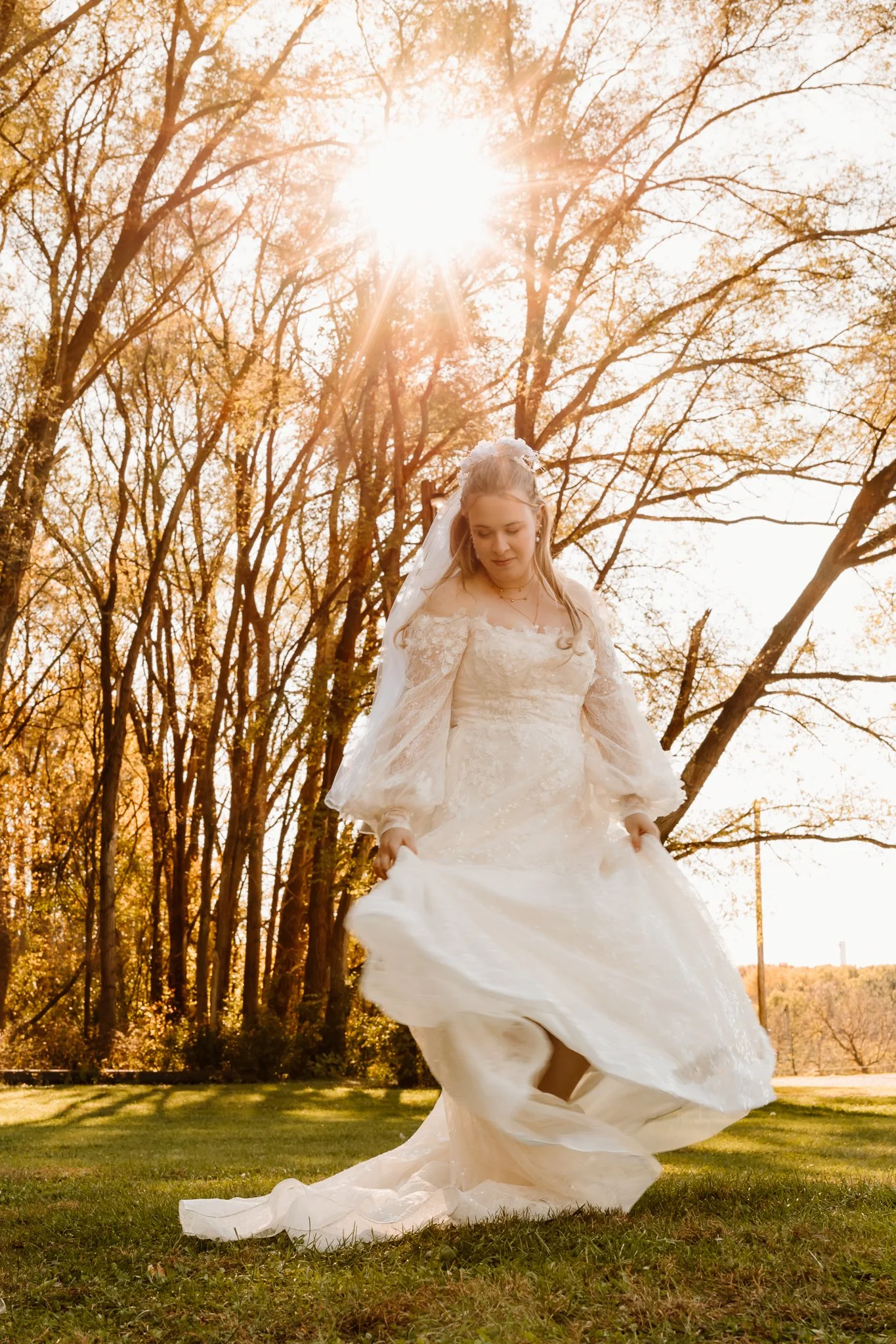 A bride in a white wedding dress walking outdoors on a grassy field with trees and sunlight in the background.