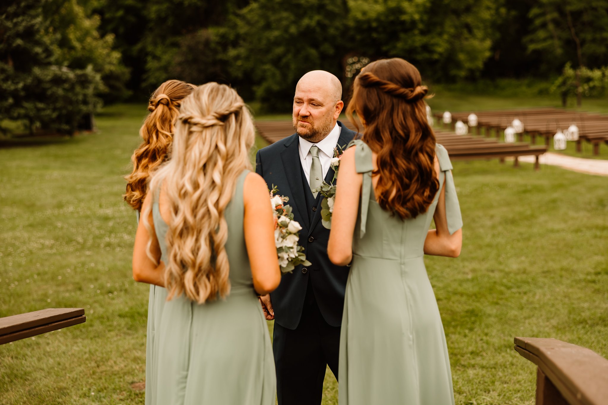 A man in a suit standing outdoors with four women in matching sage green dresses, engaged in a conversation, with a grassy area and wooden benches in the background.