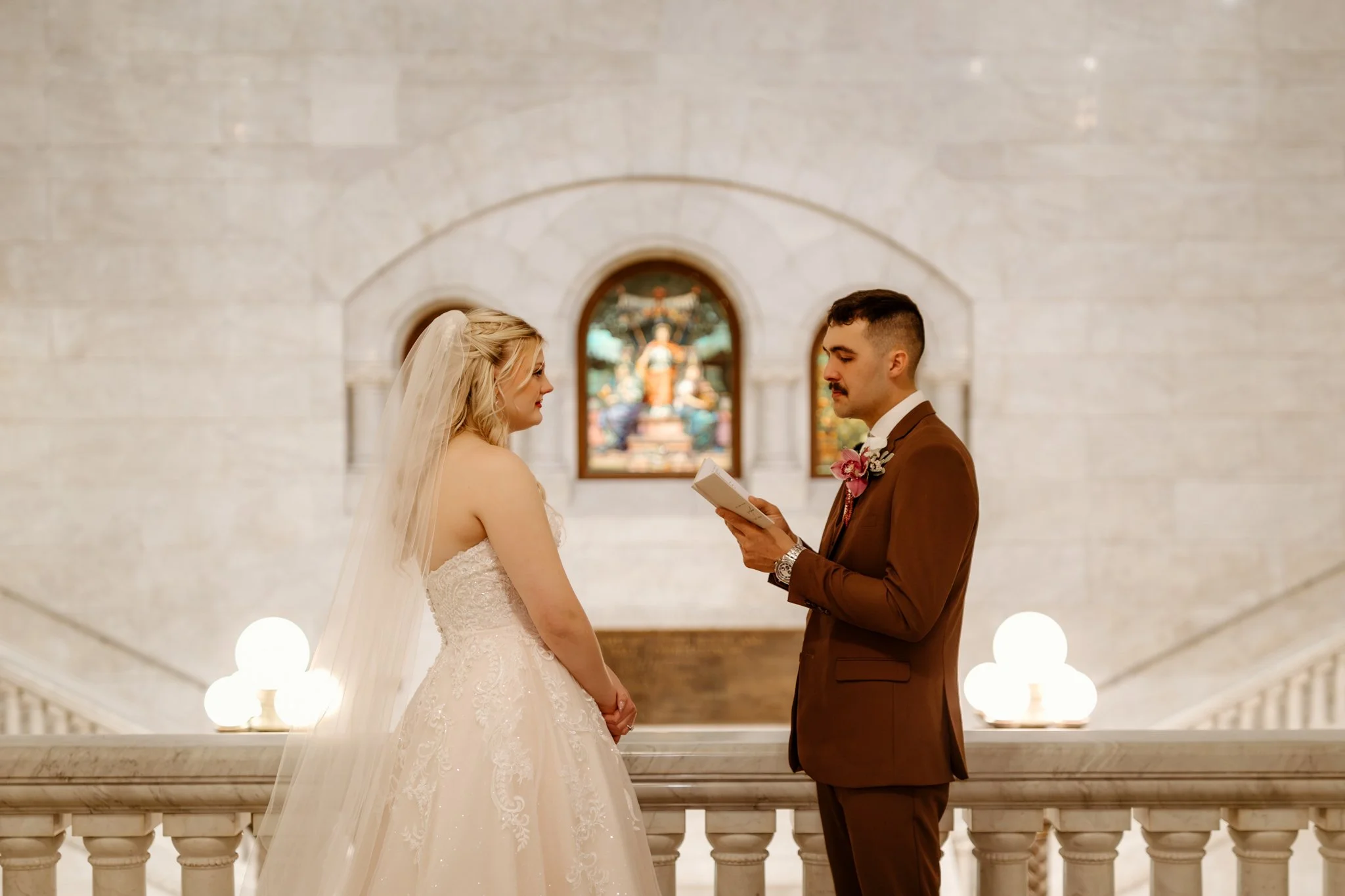 A bride and groom facing each other during a wedding ceremony inside a church. The groom is reading vows from a book, and the bride is listening. The background features stained glass windows and stone walls.