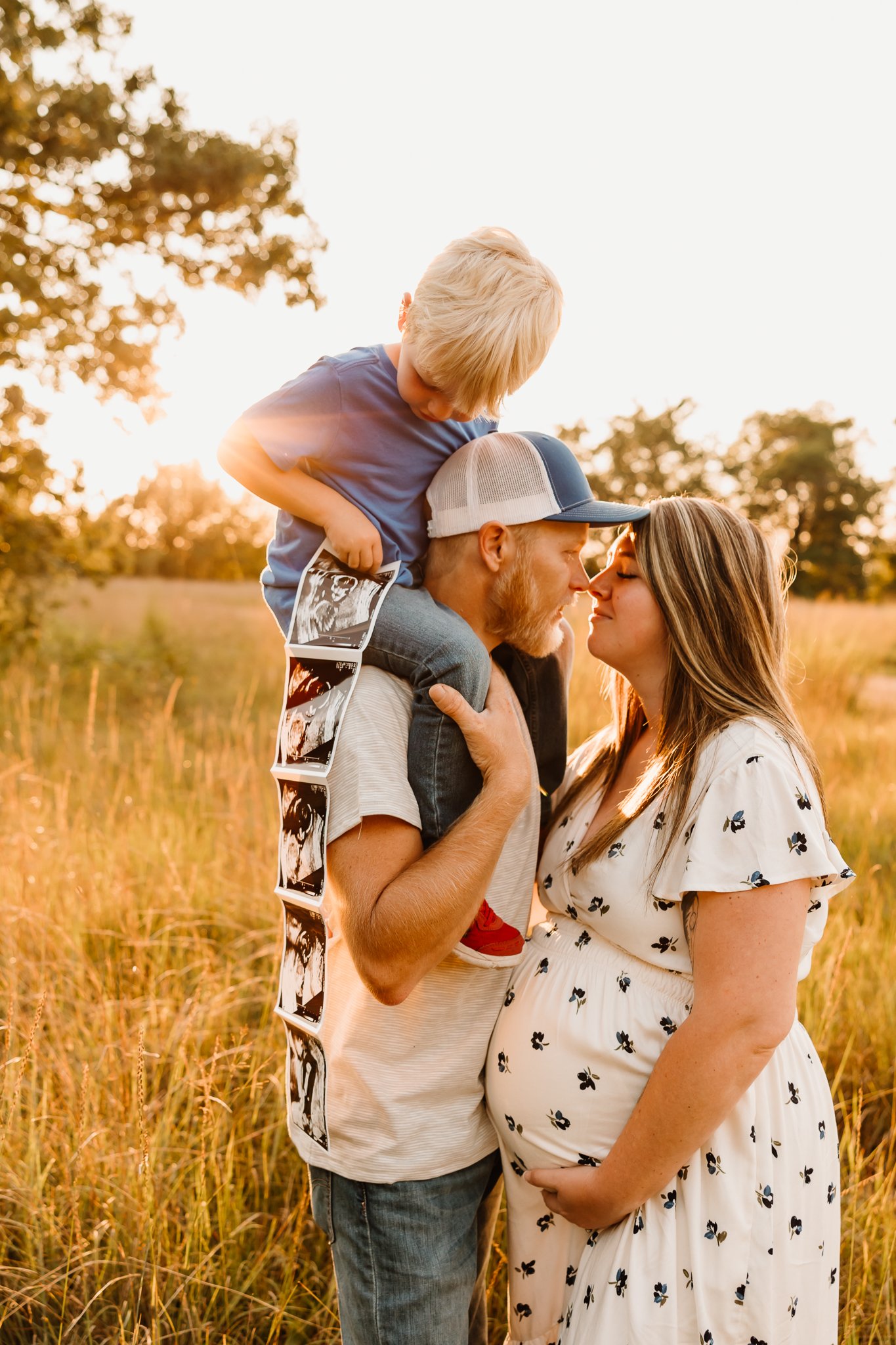 A couple, expecting a baby, sharing a tender moment in a field at sunset, with a young boy on the man's shoulders holding ultrasound images.