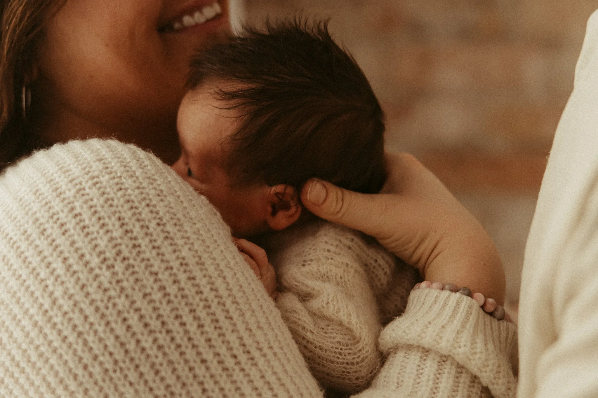 Close-up of a woman holding a sleeping baby close to her chest, both dressed in cream-colored knit sweaters, with the woman's face partially visible, smiling gently.