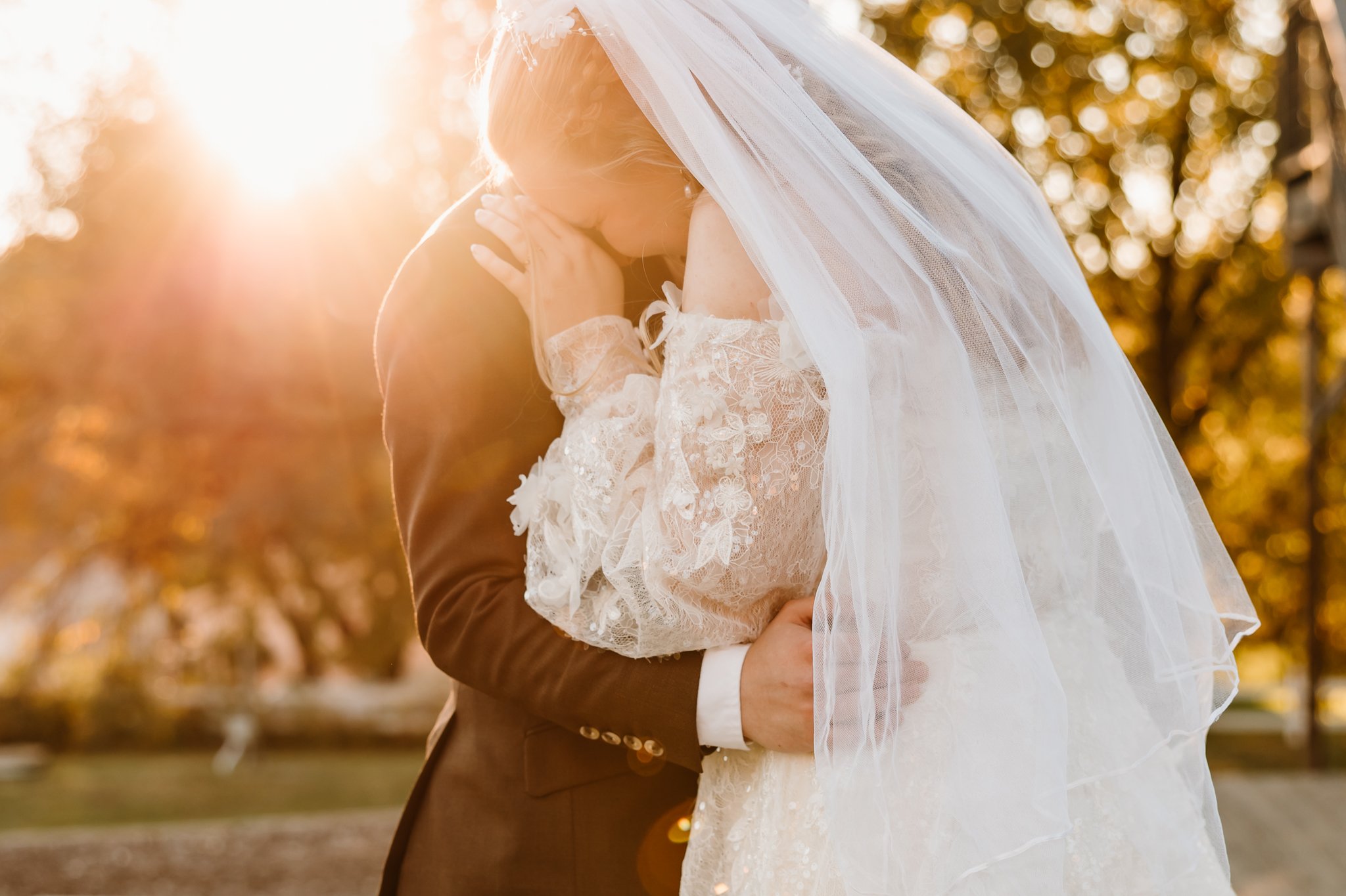 A bride and groom embracing outdoors during sunset, with the bride wearing a lace wedding dress and veil, and the groom in a brown suit.