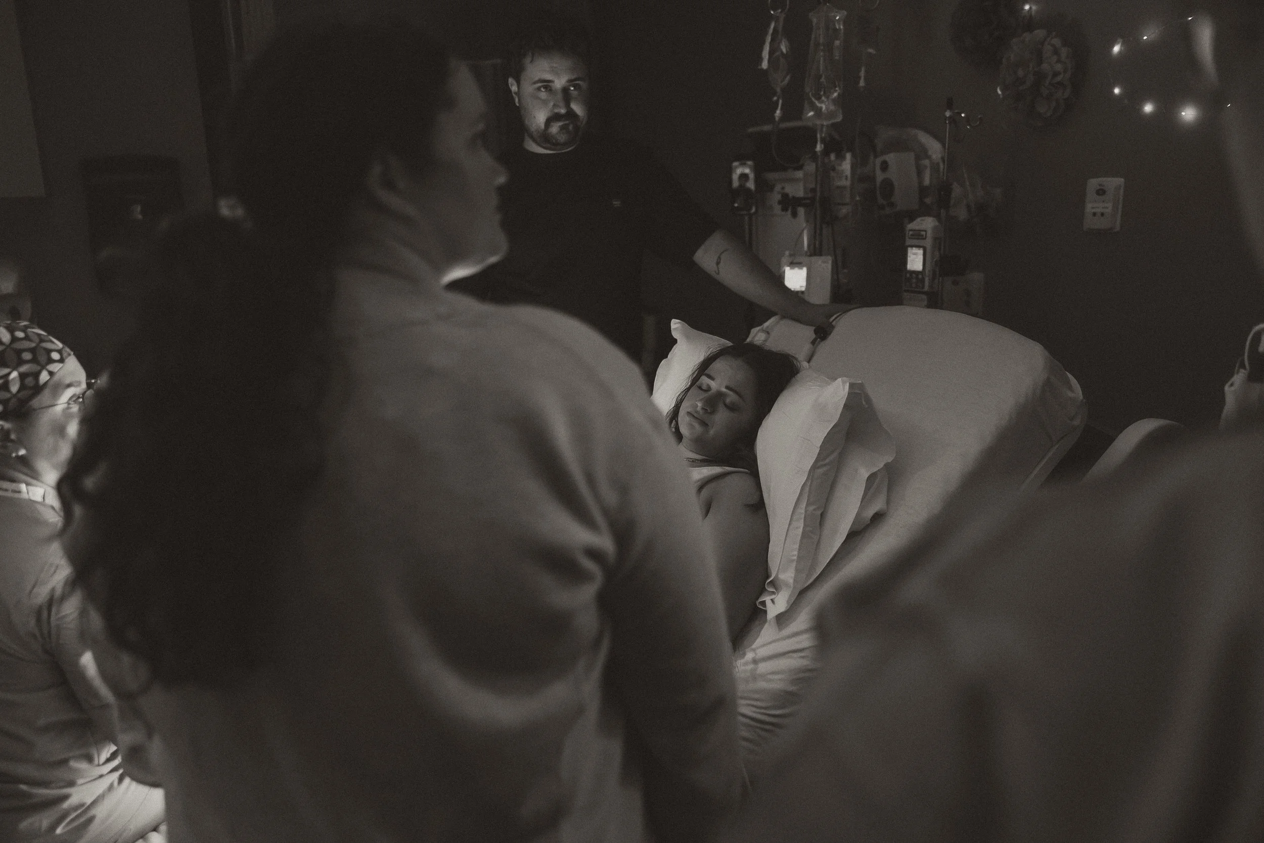 A woman lying in a hospital bed surrounded by family members, with medical equipment and bottles in the background. The photo is in black and white.