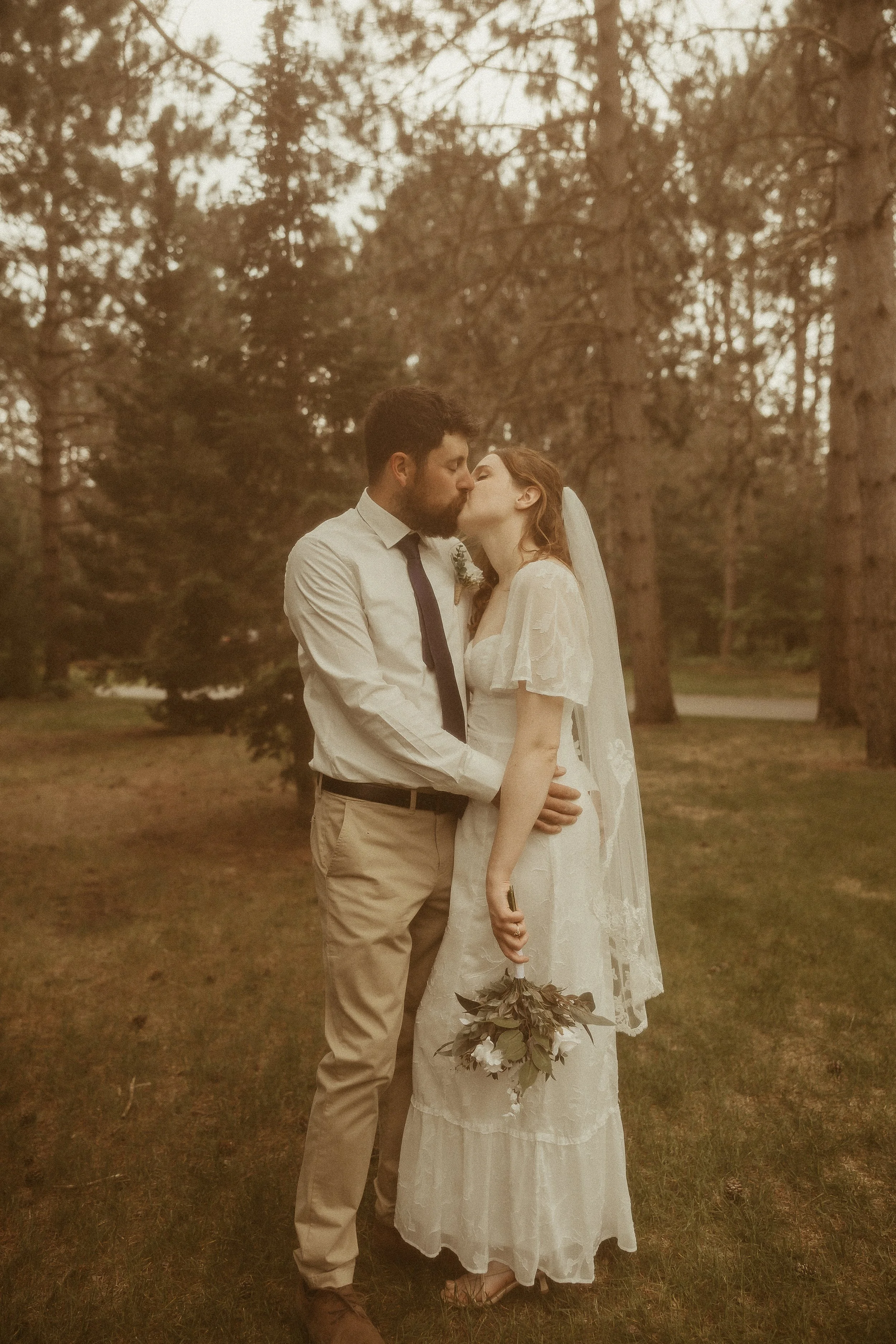 A newlywed couple sharing a kiss outdoors in a wooded area, with the woman holding a bouquet of flowers and wearing a lace wedding dress, and the man dressed in a white shirt and beige pants.