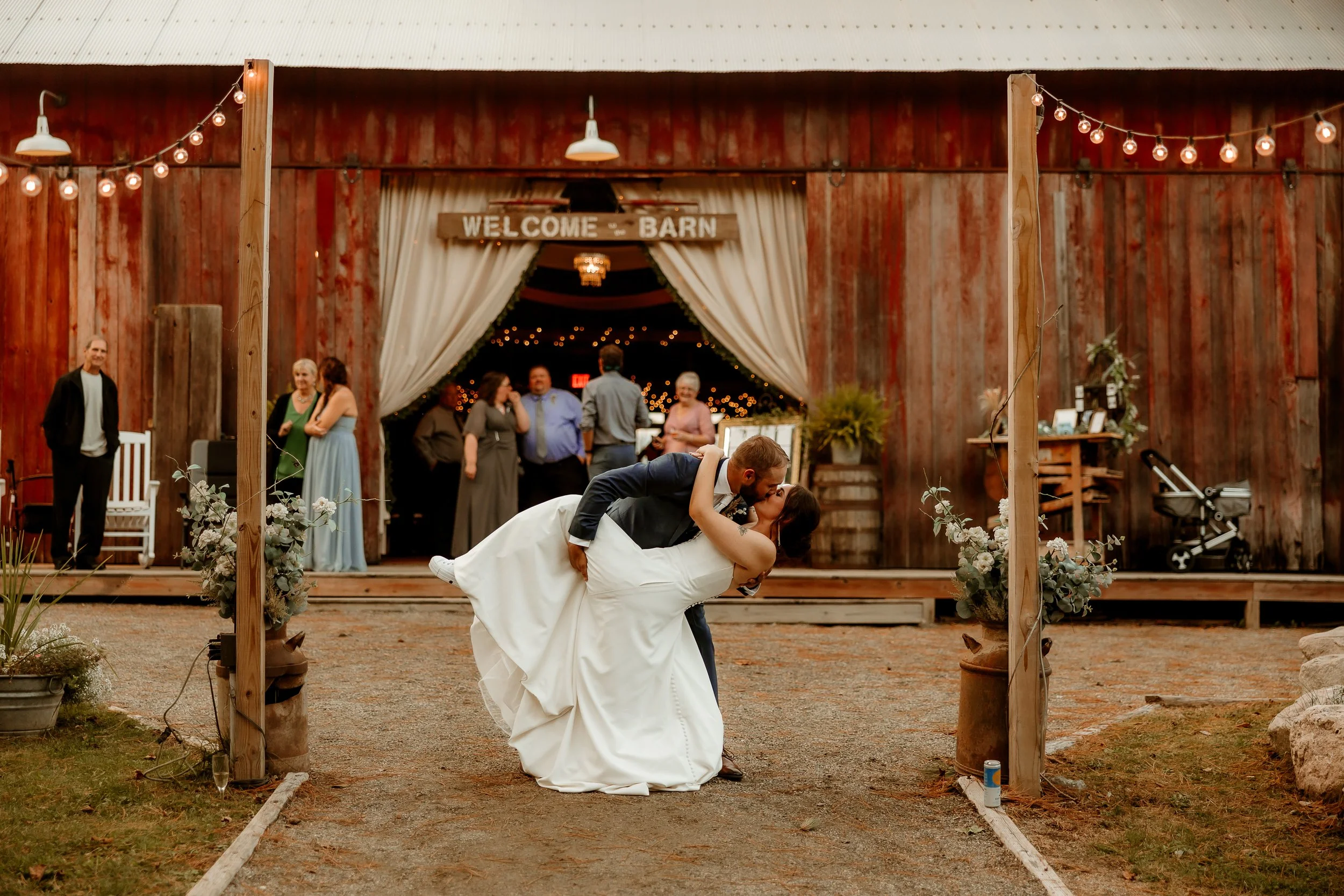 A newlywed couple dances in front of a rustic barn decorated for a wedding, with a man dipping a woman during their dance, as guests watch in the background.
