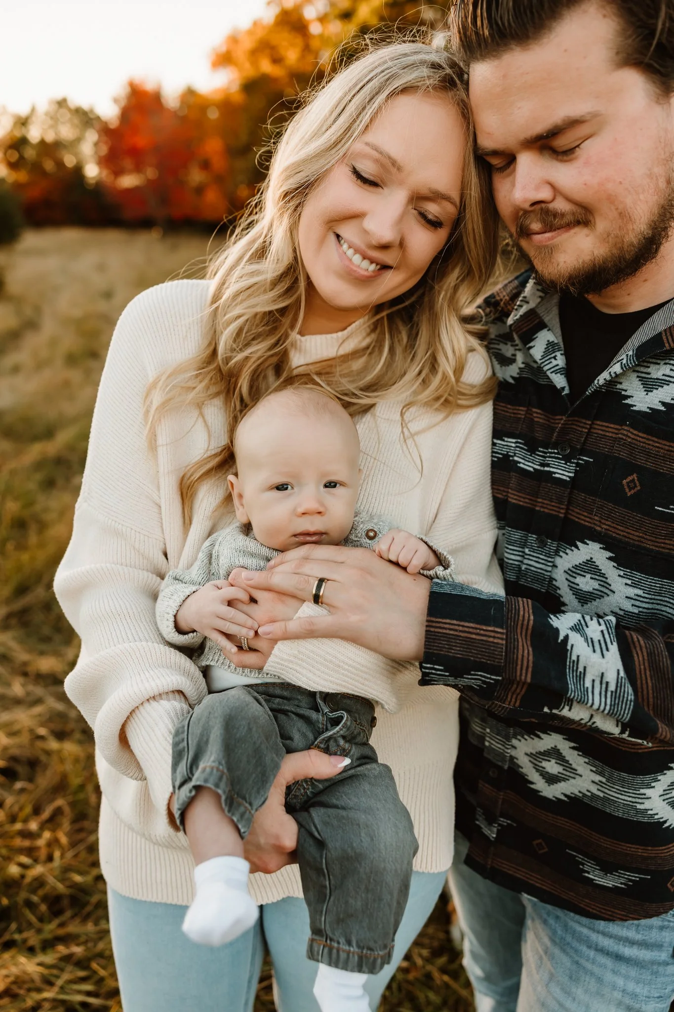 A family of three outdoors during autumn, with trees with orange and red leaves in the background. The mother, with blonde hair, is smiling and holding a baby. The father, with brown hair and a beard, is standing close and smiling with their heads to