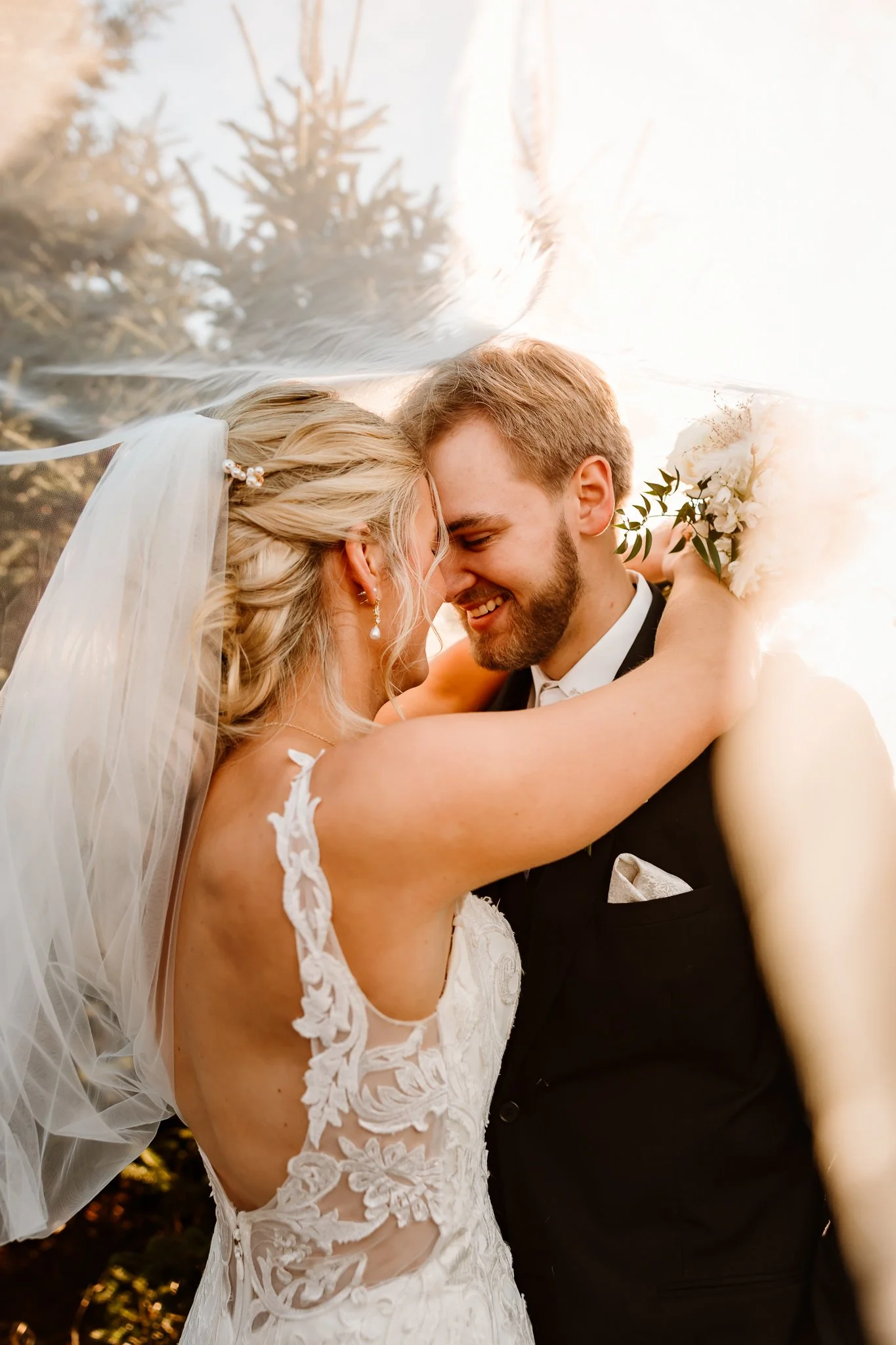 A bride and groom sharing a close, joyful moment at their wedding, with the bride wearing a lace wedding gown and veil, and the groom in a black tuxedo holding a bouquet of white flowers.
