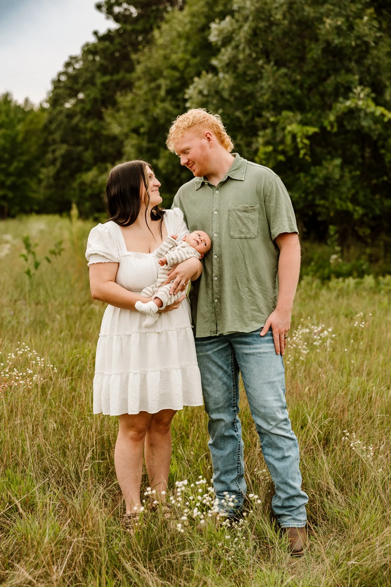A family of three standing in a field of tall grass with trees in the background, the mother holding a newborn baby, while the father looks down at the mother and child.