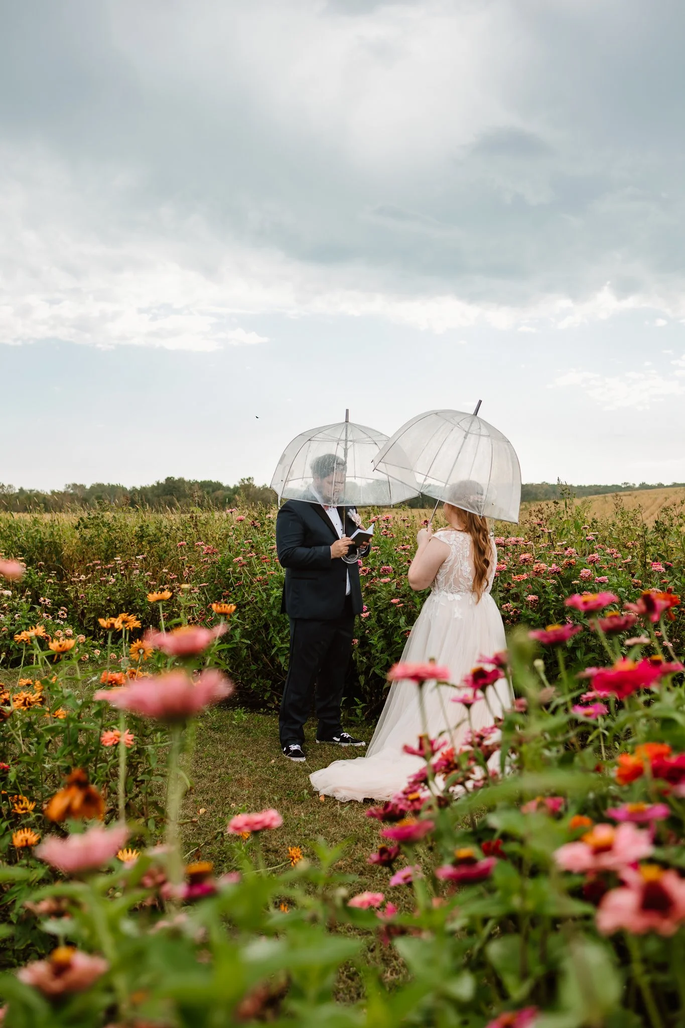 A bride and groom standing outdoors in a flower field, holding clear umbrellas, with the groom reading from a book during wedding vowsCambridge Pine city Minnesota 