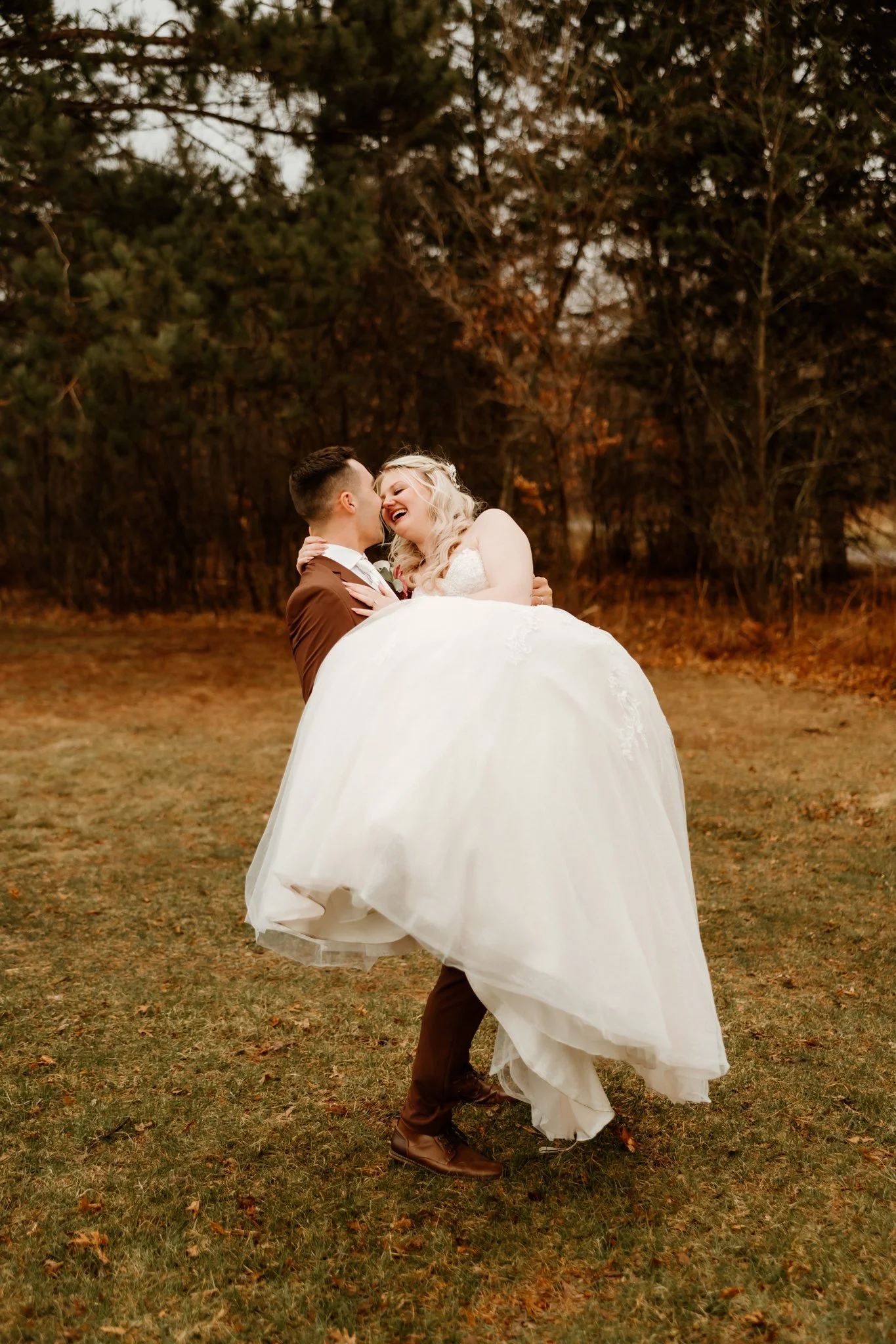 A groom lifting a bride in a white wedding dress outdoors with trees in the background, laughing and sharing a joyful moment.