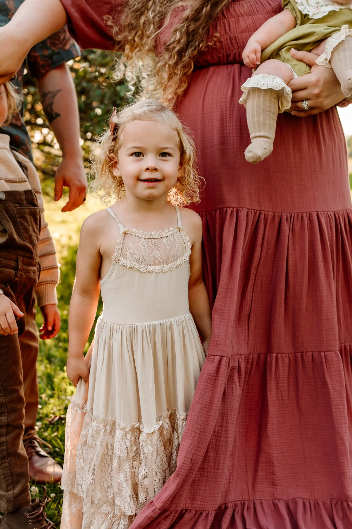 A young girl with curly blonde hair standing outdoors, wearing a cream-colored dress with lace details. She is surrounded by adults, one holding a baby, in a natural outdoor setting with sunlight.