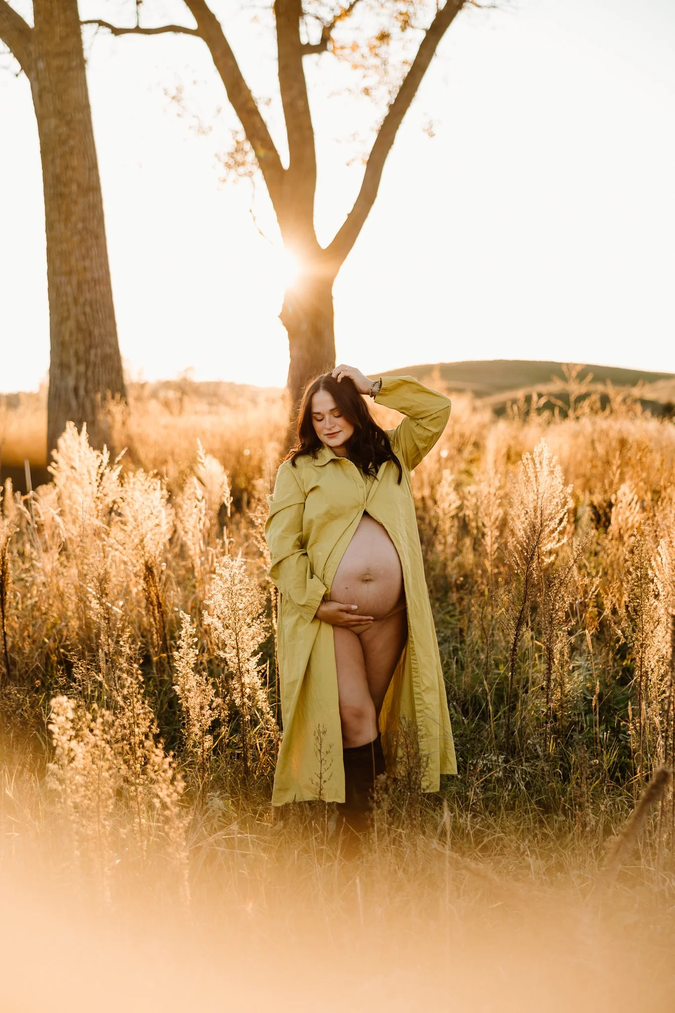 A pregnant woman in a yellow trench coat standing in a grassy field during sunset, with trees in the background.