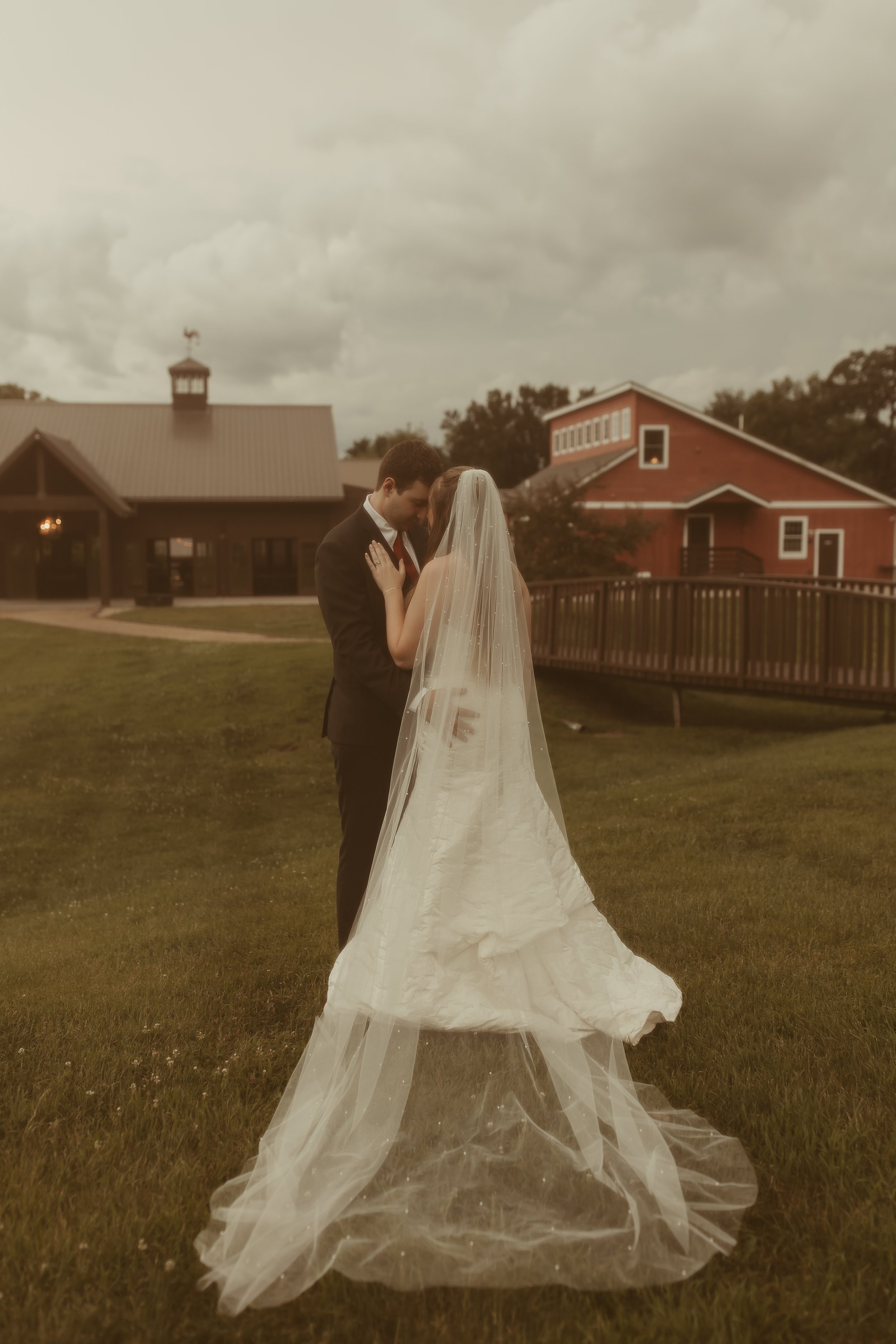 A bride and groom share a kiss outdoors on a cloudy day with a barn and a building in the background.
