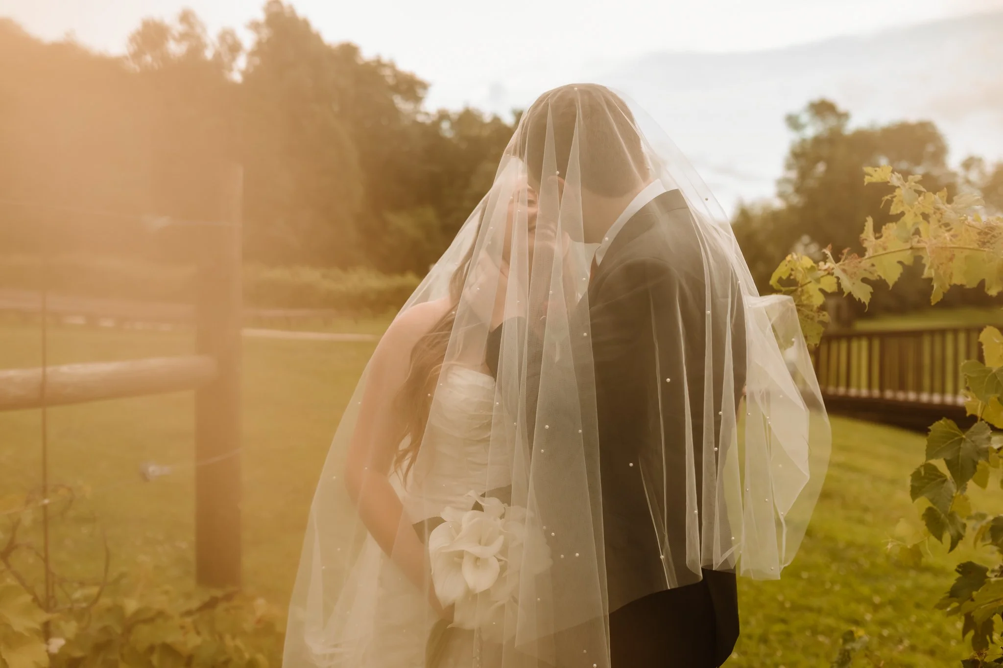 A bride and groom face each other under a bridal veil outdoors on a grassy area with trees and a wooden fence in the background.