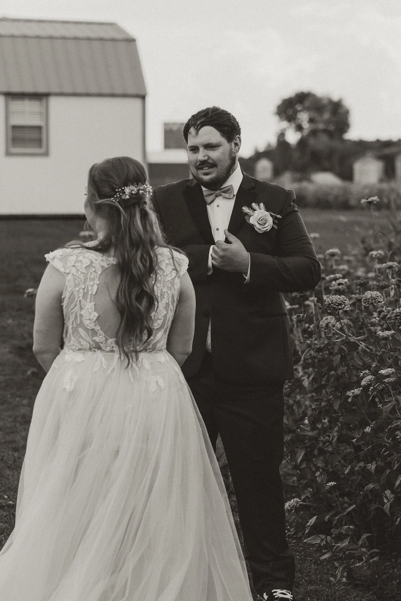 A black and white photo of a bride and groom outdoors, with the groom talking to the bride. The bride is wearing a wedding dress with lace details on the back, and the groom is in a tuxedo with a bow tie and boutonniere. There are flowers and a build
