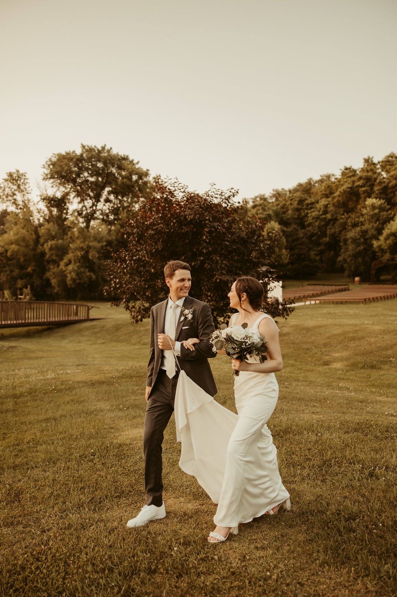 A bride and groom walking in a park during sunset, smiling at each other. The bride is holding a bouquet of flowers, and the groom is dressed in a suit with sneakers.
