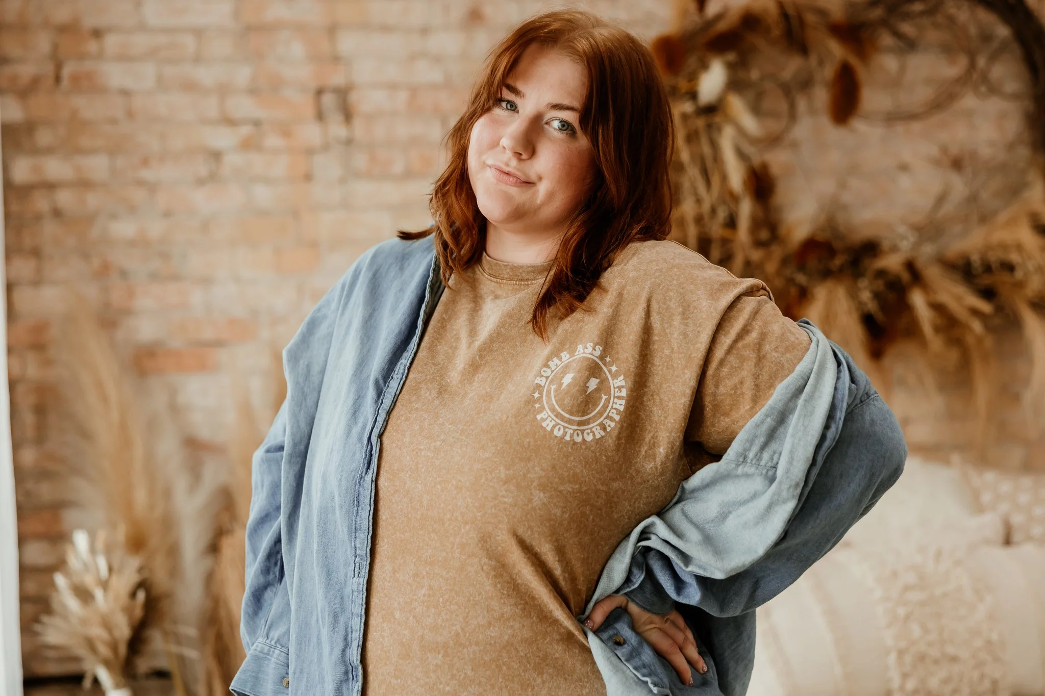 A young woman with red hair and blue eyes standing indoors, wearing a brown t-shirt with a smiley face and lightning bolts, and a denim jacket draped over her shoulders. She has one hand on her hip and is posing casually.