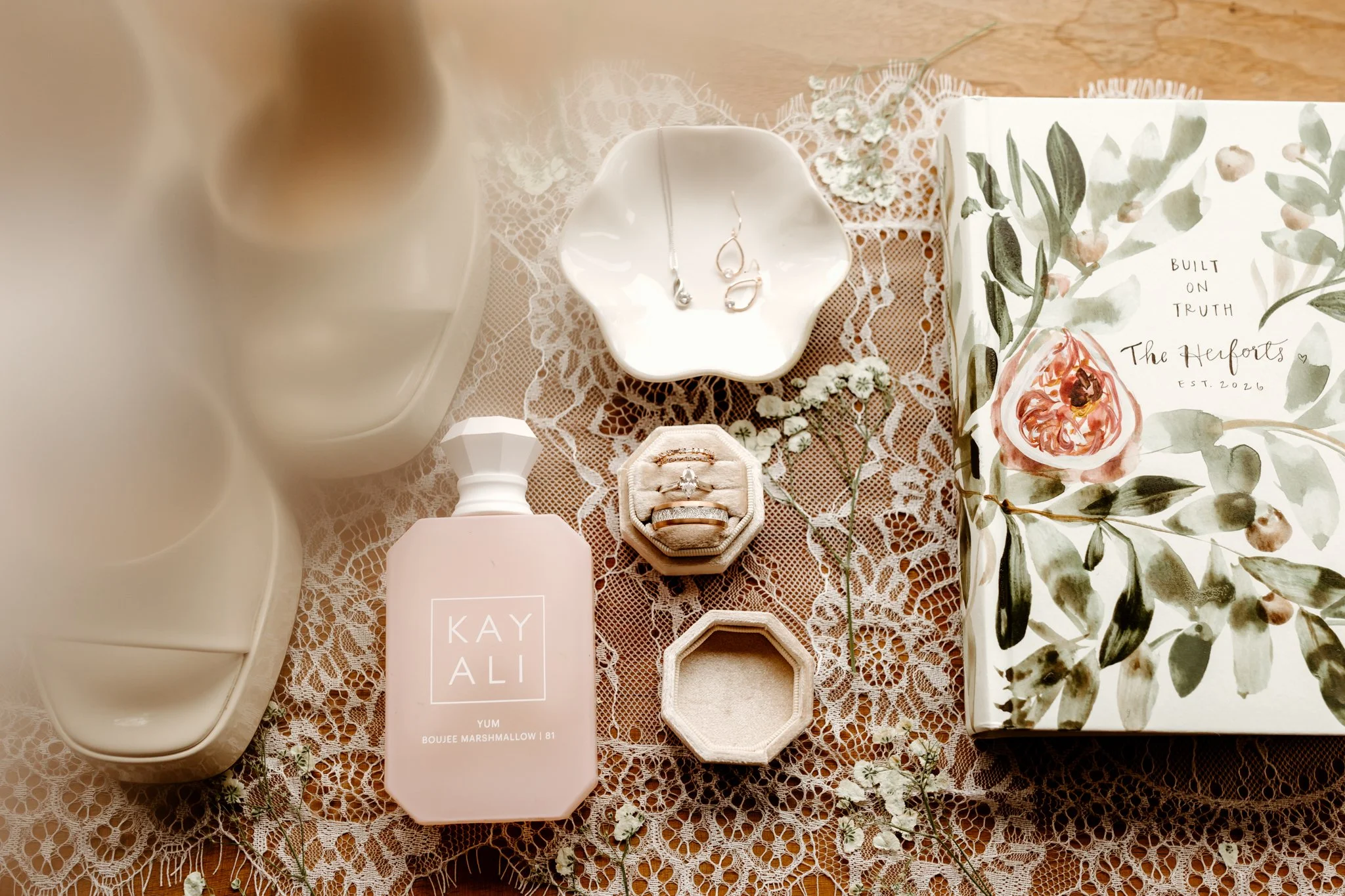 A collection of jewelry, a perfume bottle, a decorative box, and a floral book placed on lace fabric. The jewelry includes earrings and rings, with the perfume bottle labeled 'KAYALI Yum Boujee Marshmallow.' The decorative box is open and empty. The 