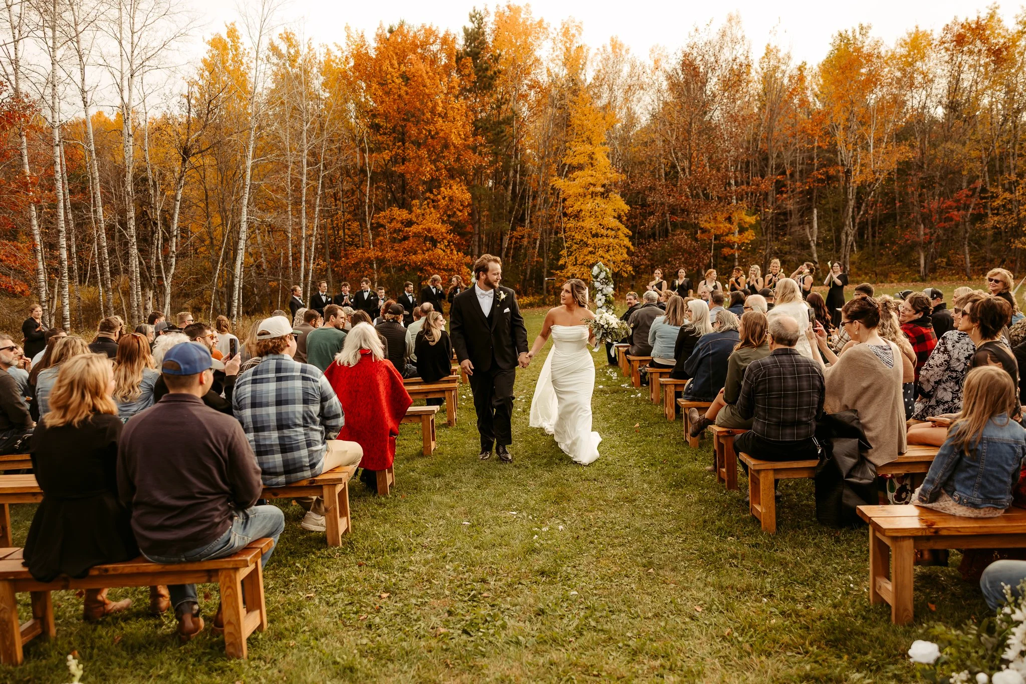 A wedding ceremony outdoors in fall with the bride and groom holding hands, surrounded by seated guests on wooden benches, and a background of colorful autumn trees.