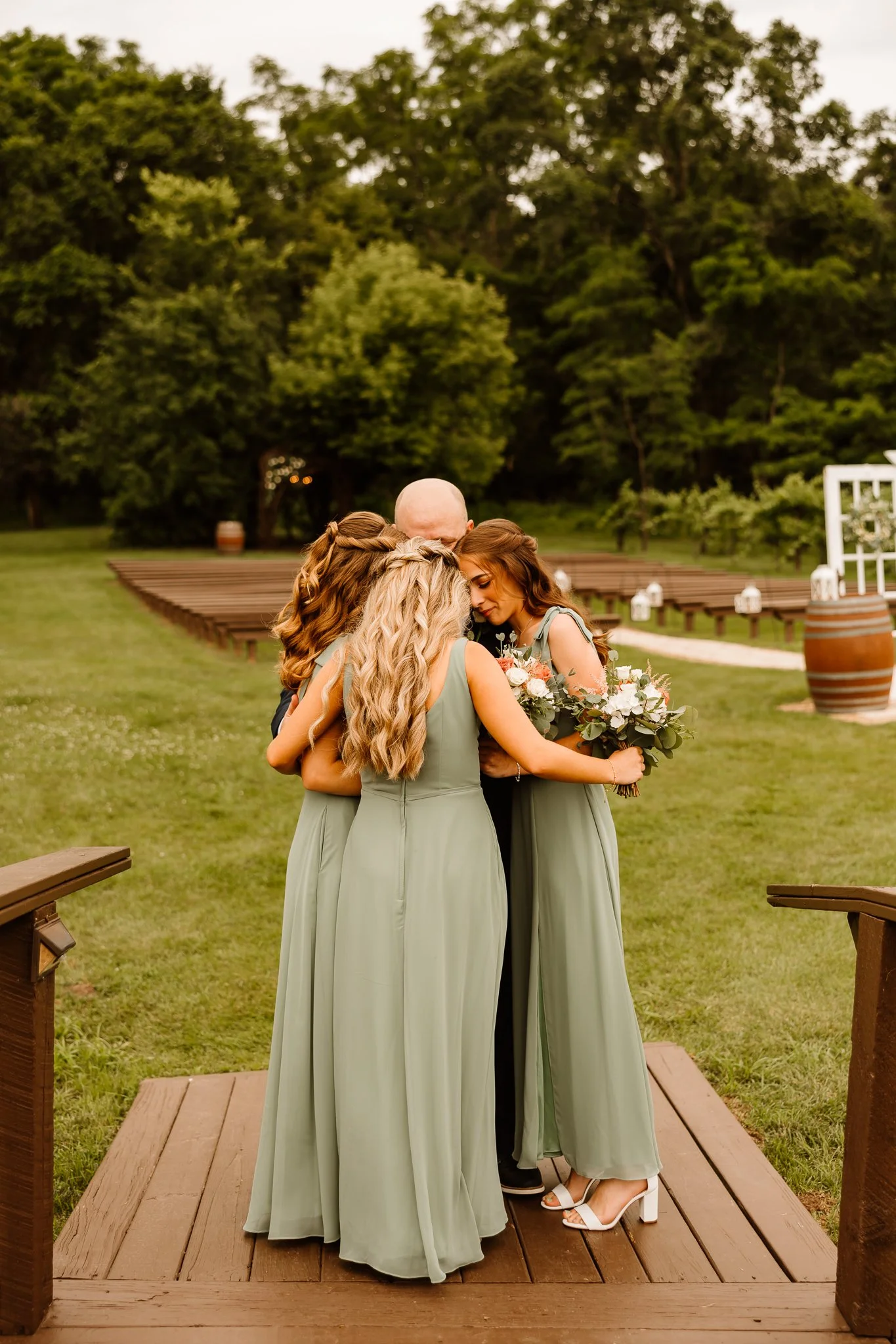 A group of women in light green dresses embracing on a wooden platform outdoors during a wedding ceremony, with trees and decorated aisle in the background.