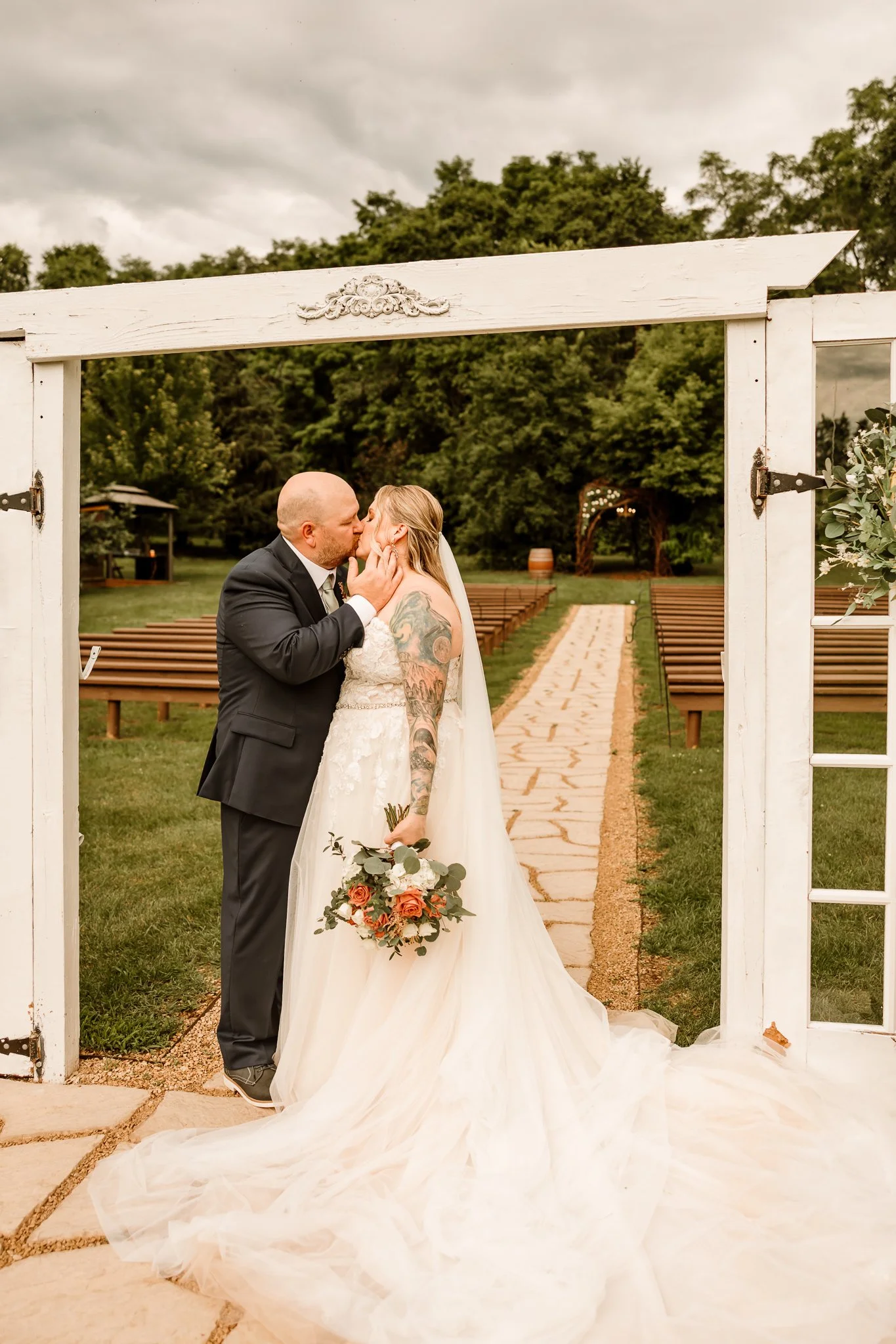 A newlywed couple sharing a kiss under a white wooden arch at an outdoor wedding ceremony, with chairs arranged on either side and a stone pathway leading to an archway in the backgroundHope Glen Farm Cottage Grove Minnesota 