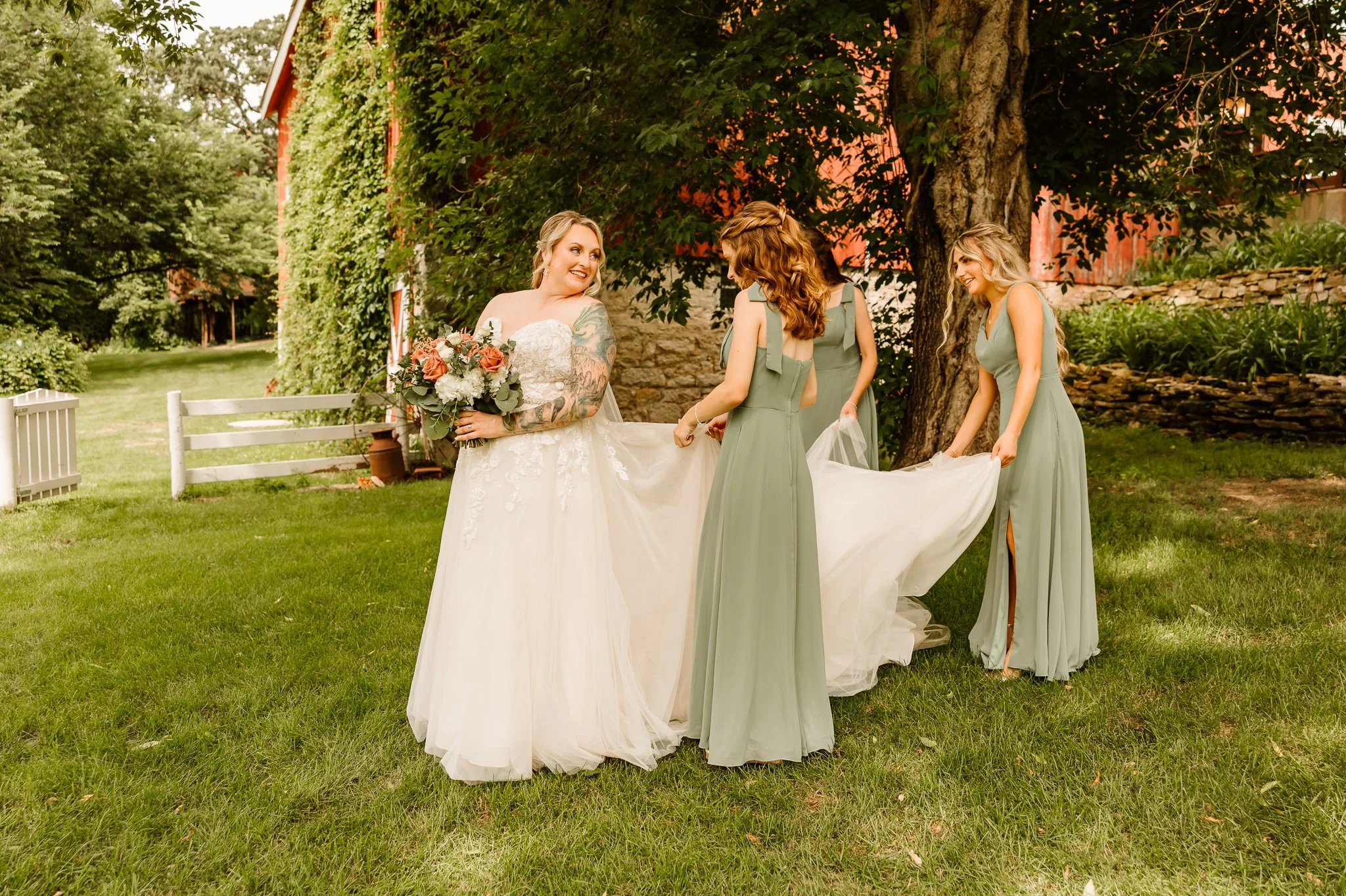 Bride in wedding dress holding bouquet standing on grass with three bridesmaids adjusting her train outdoors near trees and a rustic red building.