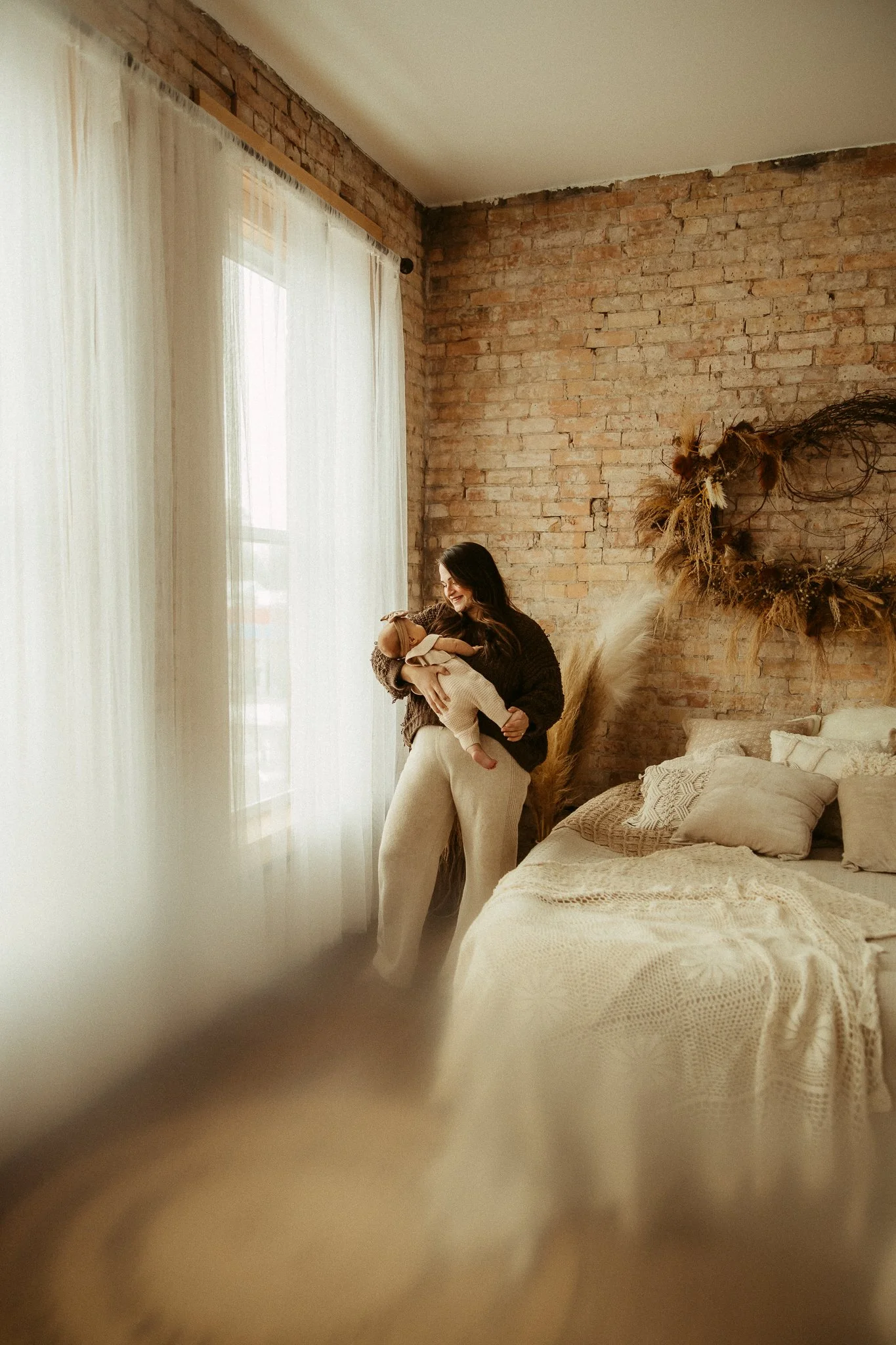 Woman holding a baby in a cozy bedroom with a brick wall, decorated with dried flowers and pampas grass, next to a window with white curtains.