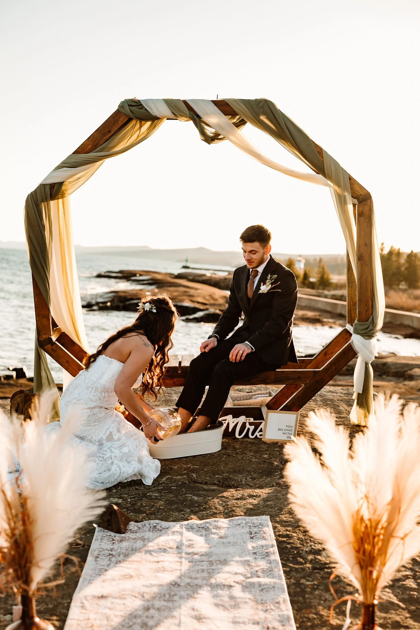 A couple gets married on a rocky beach at sunset, with the groom sitting and the bride pouring water over his feet during a ceremony under a wooden arch decorated with draped fabric, surrounded by pampas grass.