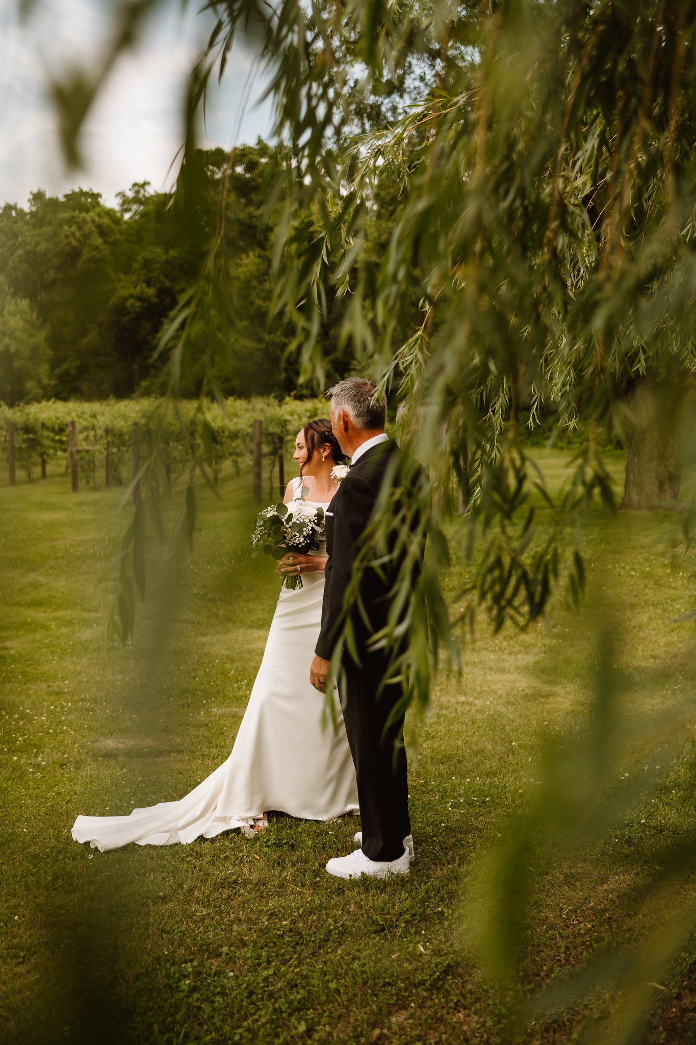 A bride in a white wedding dress holding a bouquet, standing next to a groom in a black suit with white sneakers, in an outdoor setting surrounded by greenery and trees, with leaves partially obscuring the view.