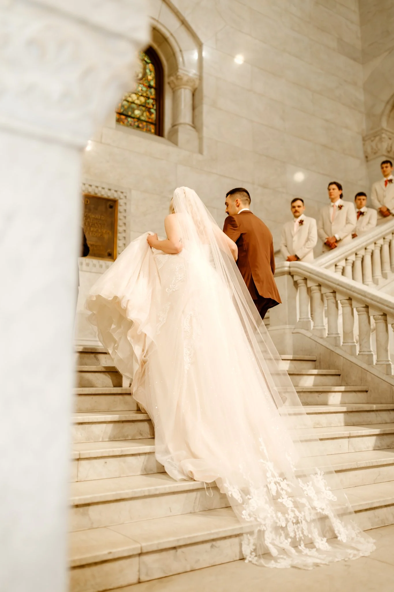 Bride in a wedding gown and veil ascending church stairs with groom and groomsmen in white suits standing nearby.