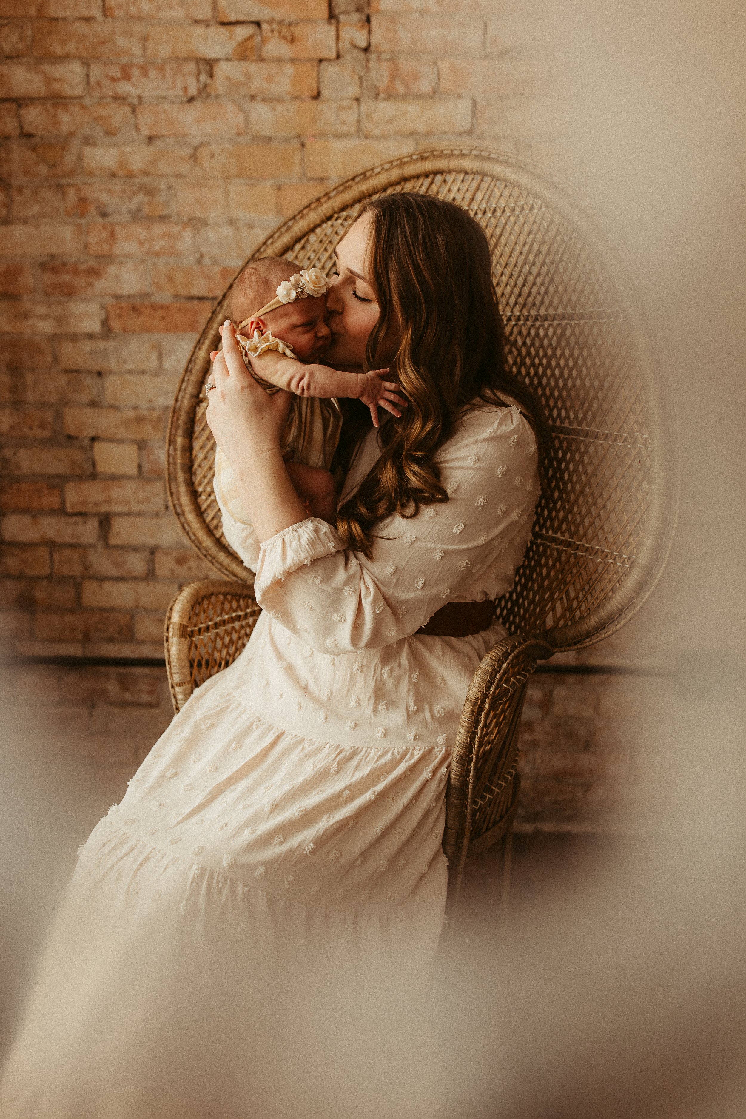 A woman sitting on a wicker chair holding and kissing a sleeping baby with a floral headband. The background is a brick wall.