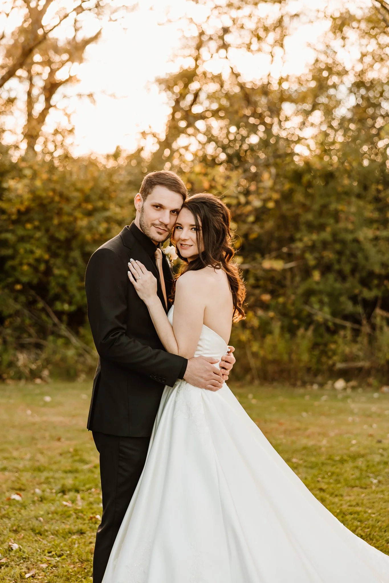 A bride and groom in wedding attire standing outdoors at sunset, embracing each other in front of a background of trees with warm, golden light.