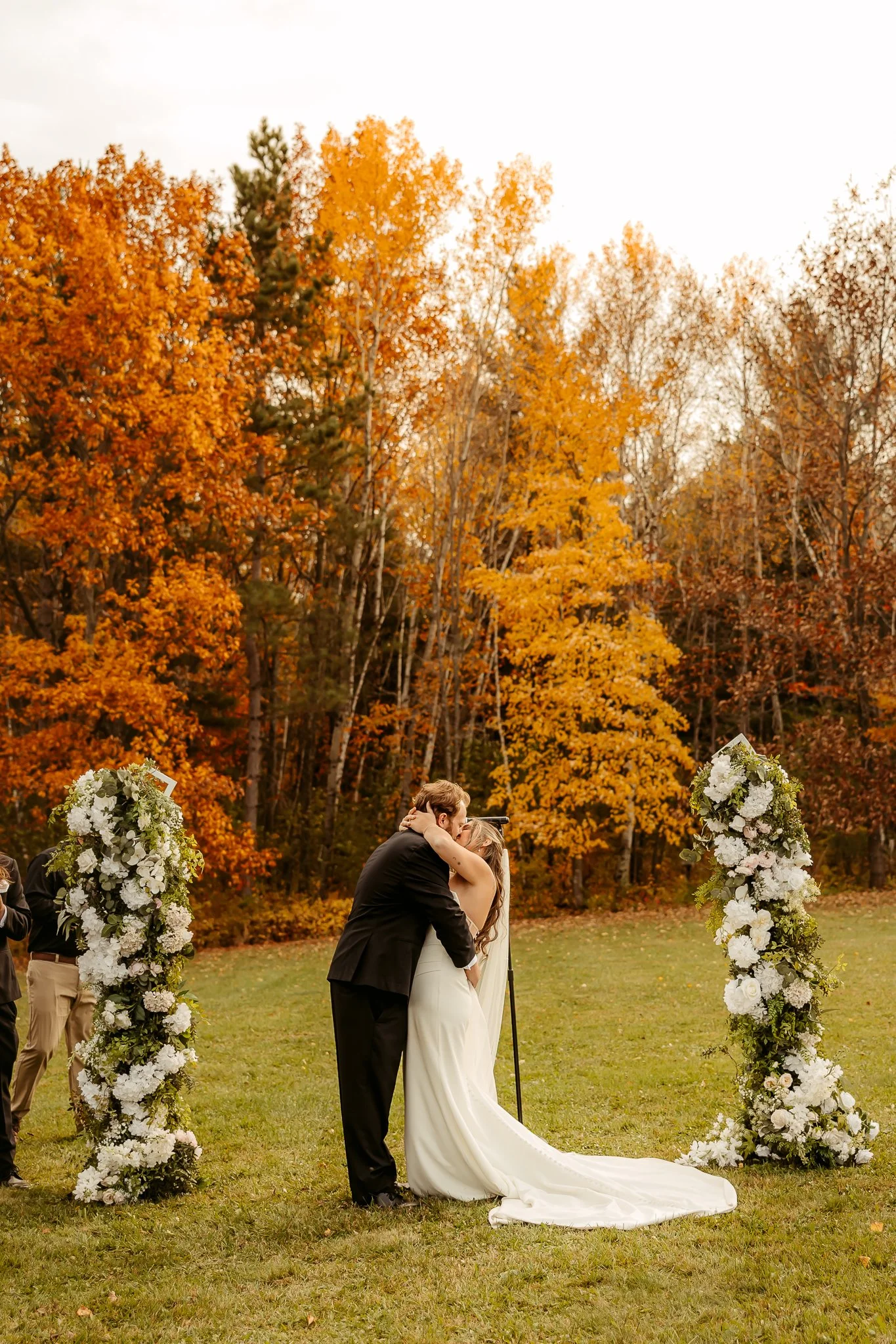 A bride and groom share a kiss during their outdoor wedding ceremony, surrounded by autumn foliage and floral arrangements.