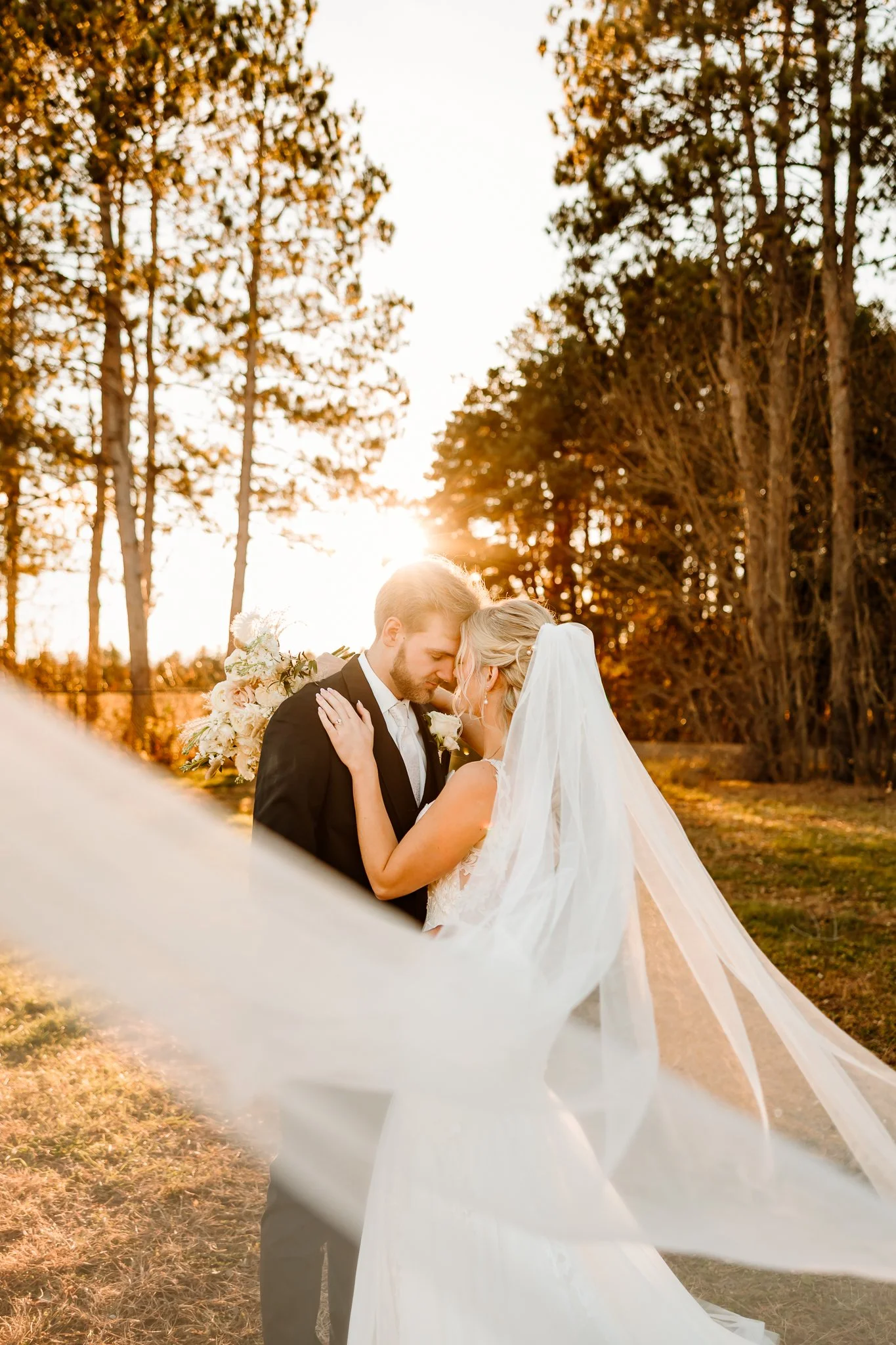 A bride and groom sharing a kiss outdoors during sunset, with the bride wearing a long veil and holding a bouquet, surrounded by trees. Cambridge Minnesota 