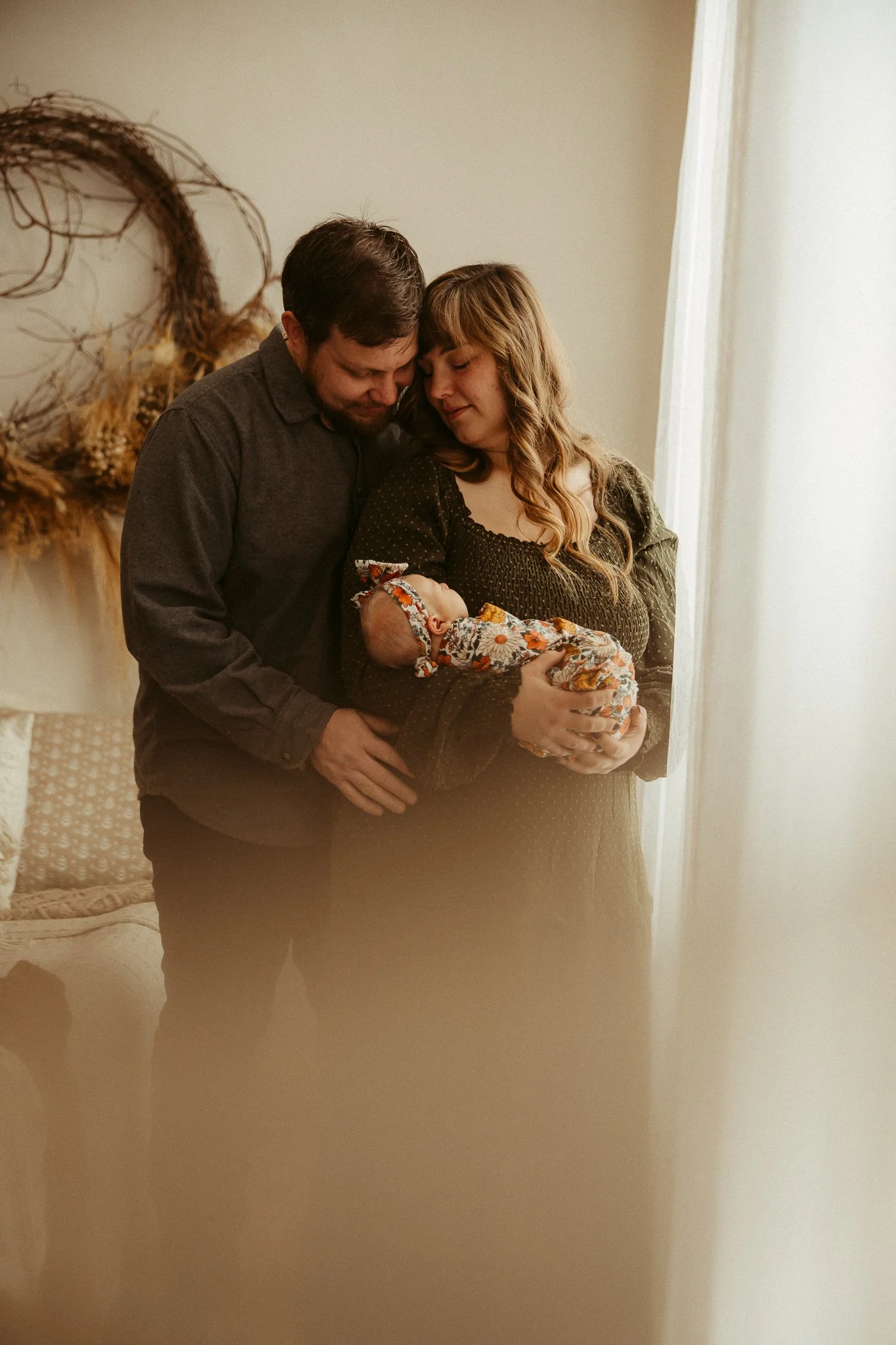 A couple holding their sleeping baby near a window, with a decorative wreath hanging on the wall behind them.