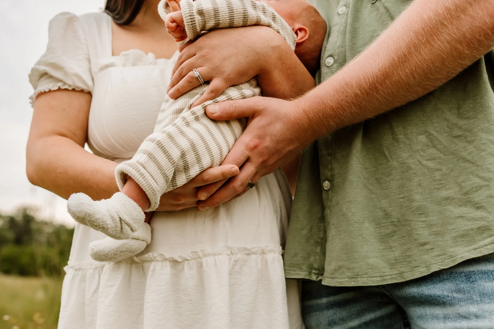 Close-up of a family holding a baby's legs outdoors, focusing on their hands and bodies, with a background of a cloudy sky and greenery.