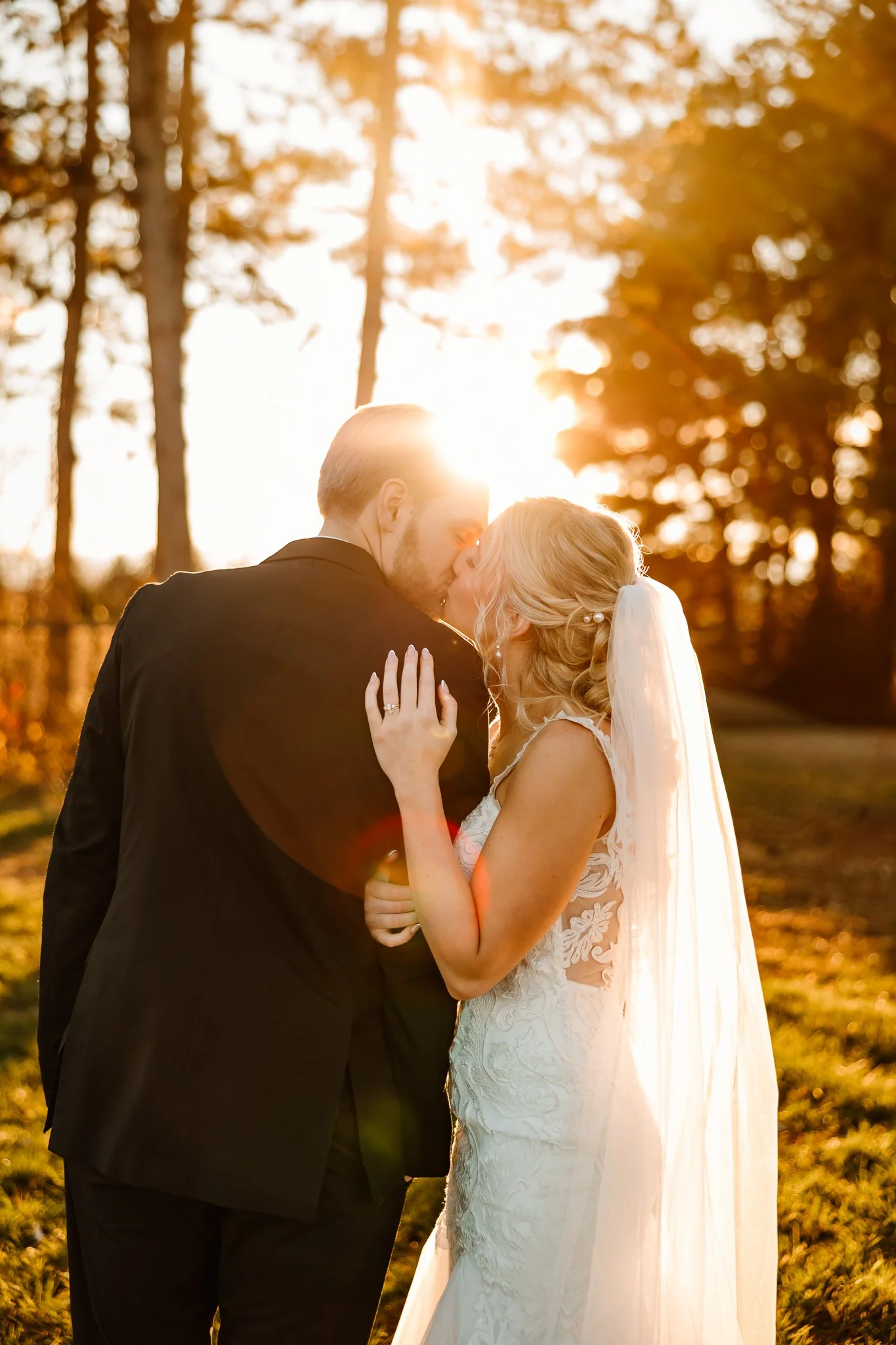 A bride and groom share a kiss outdoors during sunset, with the sun shining behind them and trees in the background.