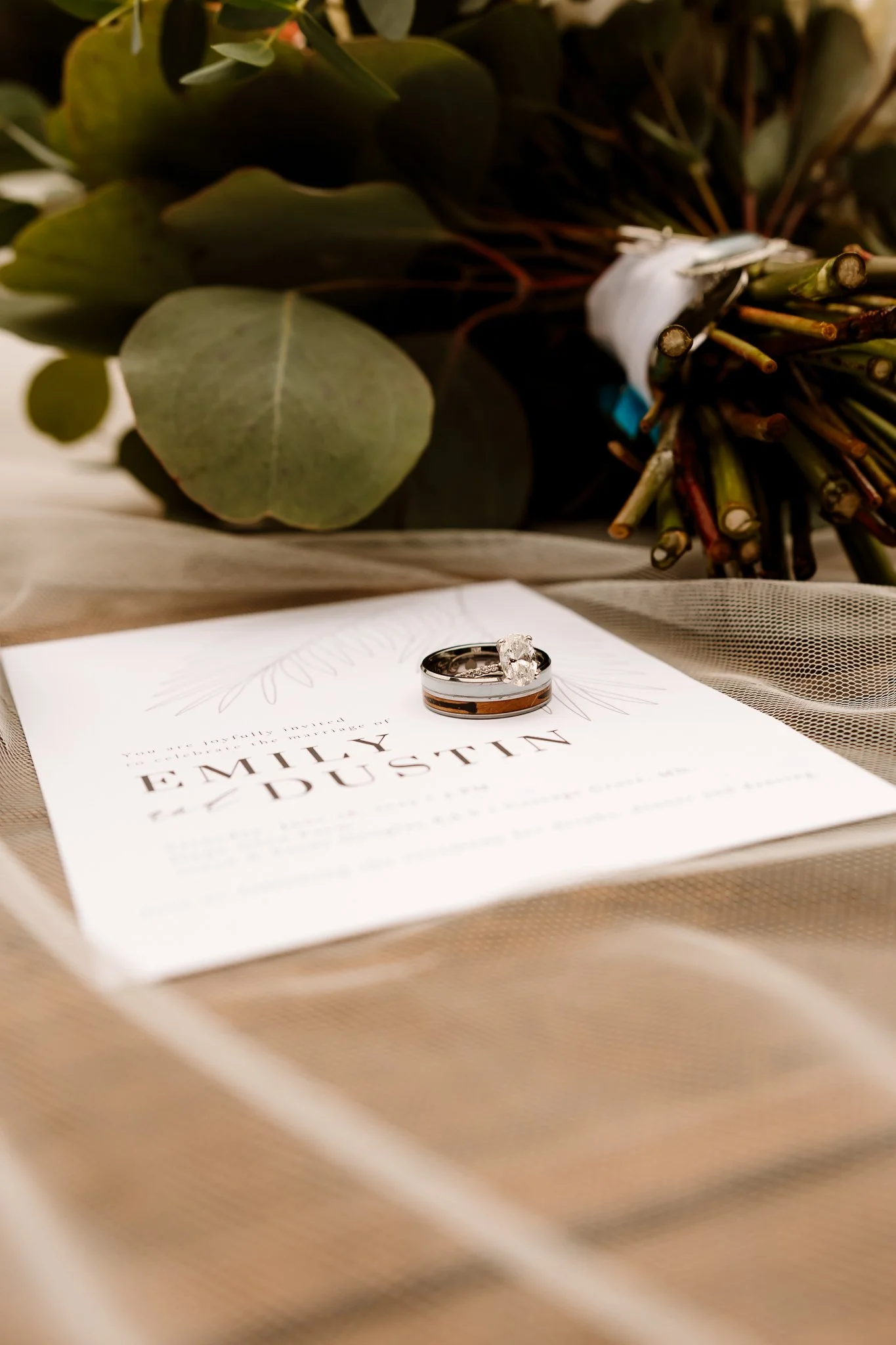 A wedding ring and engagement ring placed on a wedding program with the names Emily and Dustin, surrounded by a bouquet of flowers and greenery.