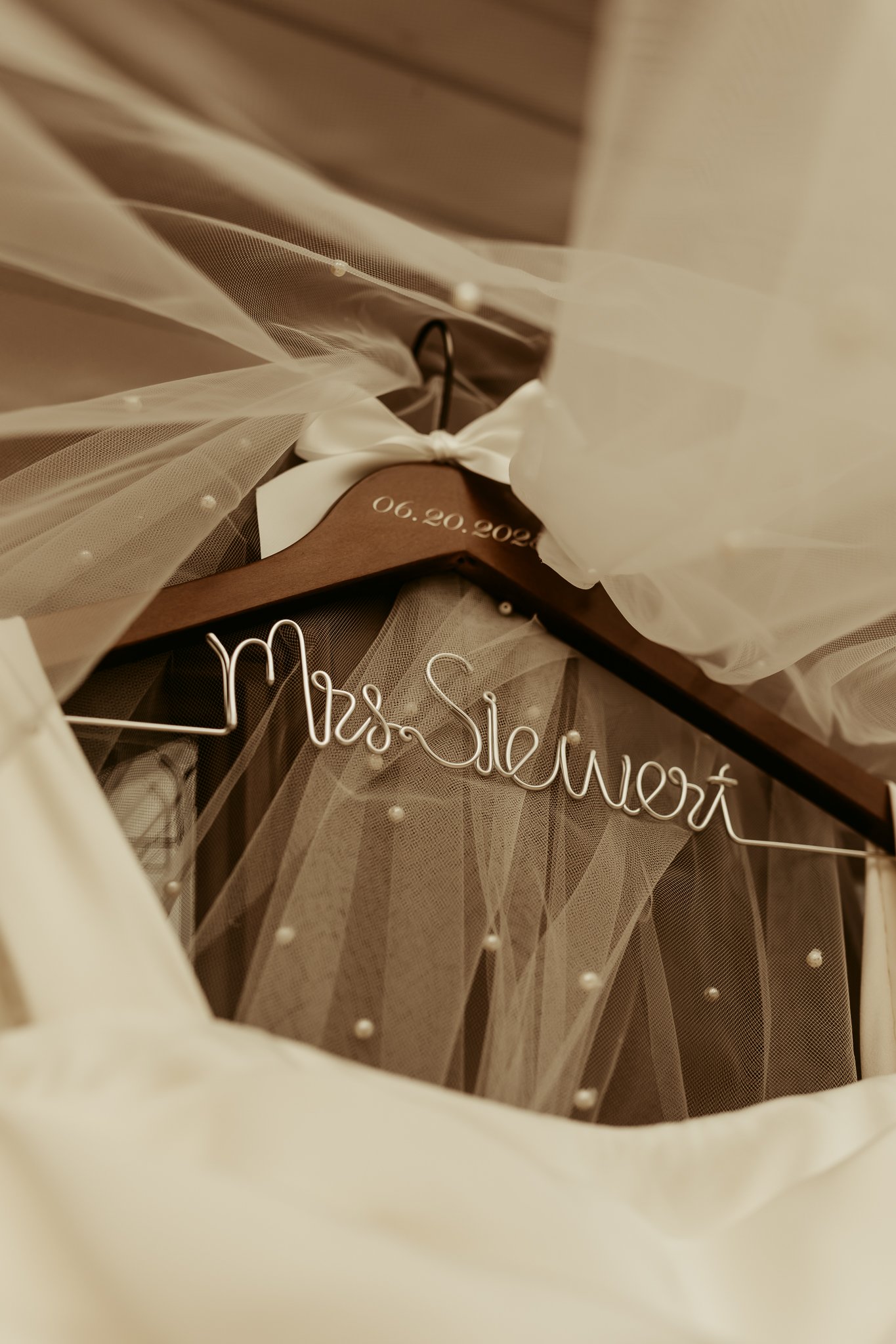 A wedding dress hanger with a metal sign reading 'Mrs. SloWert,' a wooden hanger with the date '06.20.2021,' and ivory and tulle fabric decorated with pearls.