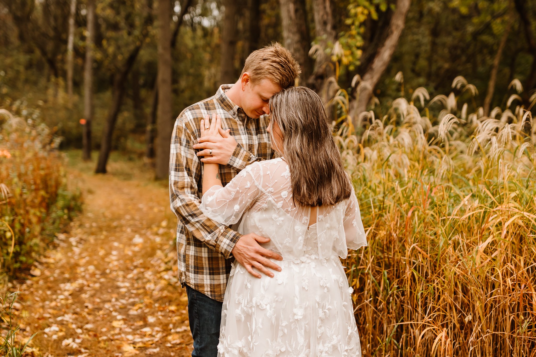 A couple holding each other lovingly in an autumnal forest, with fallen leaves on the ground and tall grasses surrounding them.