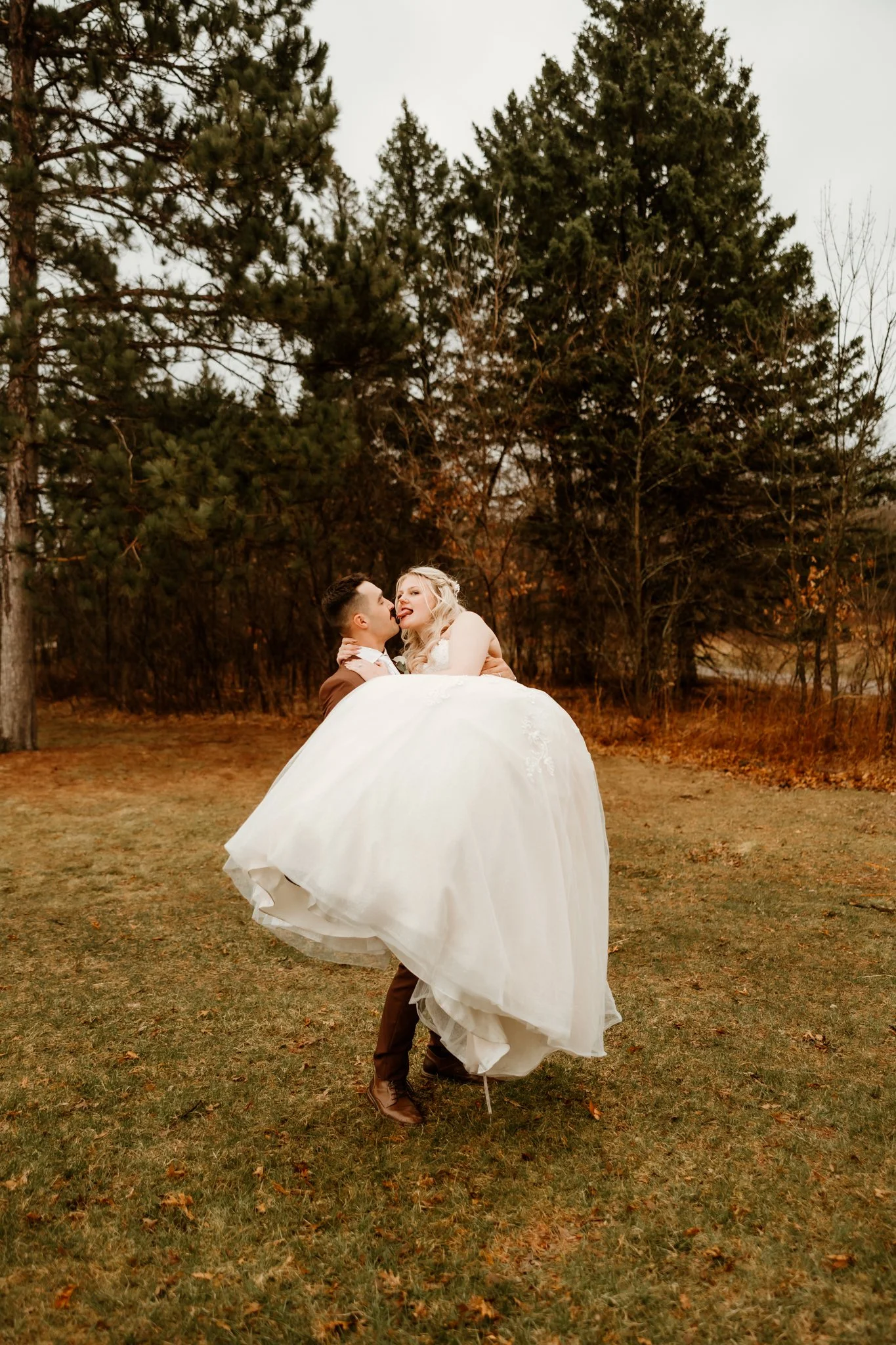 A bride and groom dancing outdoors on grass with trees in the background, the groom lifting the bride who is smiling and looking at him.