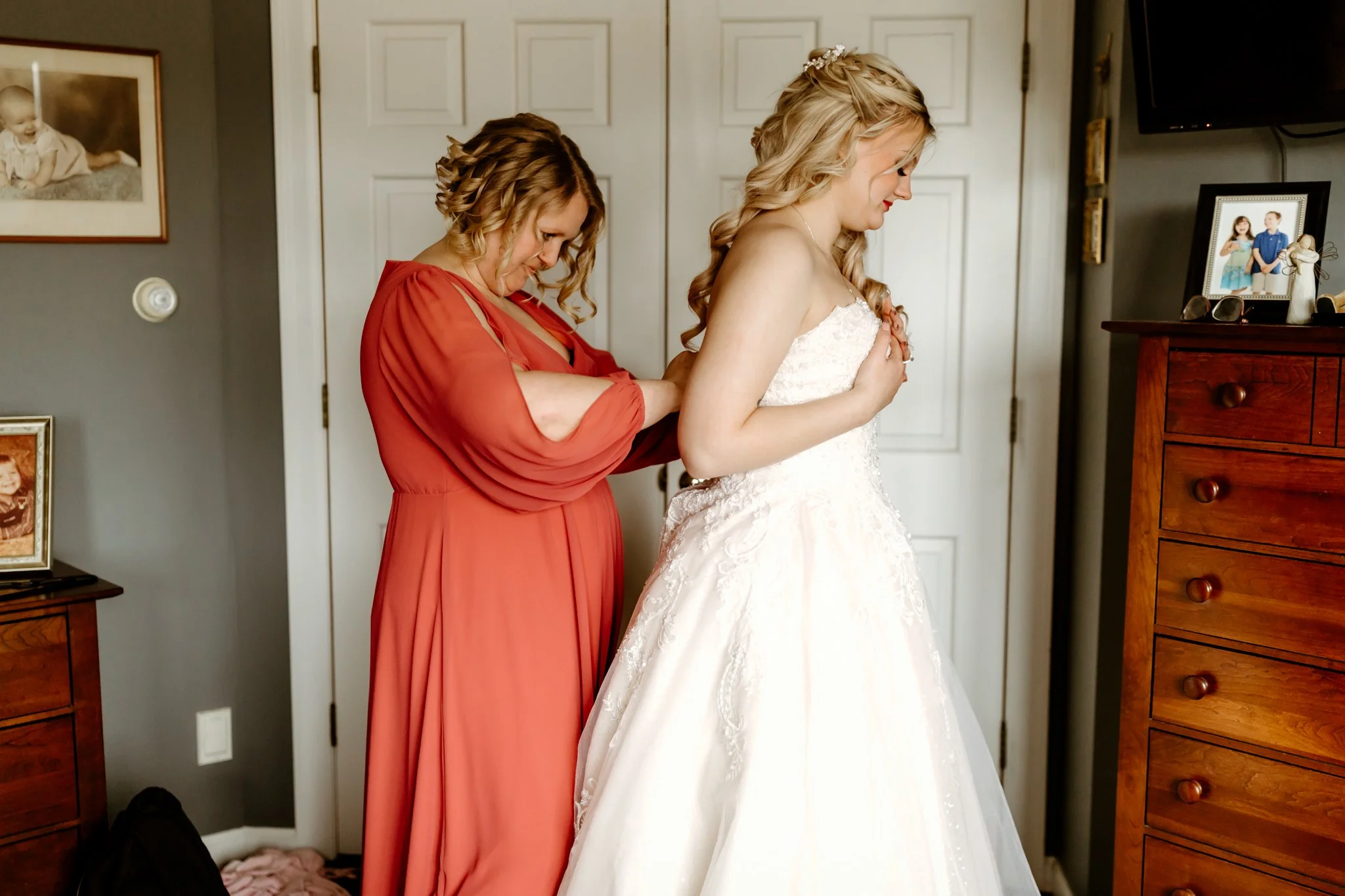 A bride in a white wedding dress with lace details is getting ready, assisted by a woman in a coral dress, in a bedroom with family photos on the dresser and wall.