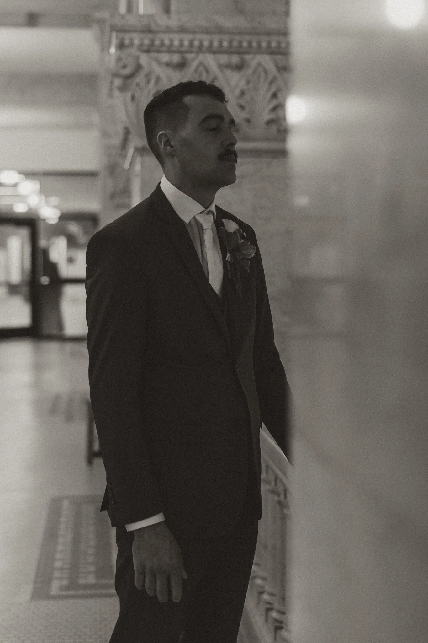 A man in a suit standing with eyes closed in a dimly lit interior with ornate architectural details.
